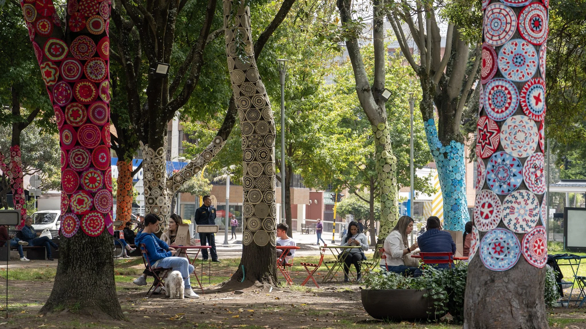 Árboles decorados con textiles coloridos, con patrones geométricos y florales, en un parque del centro de la ciudad con personas sentadas y caminando.
