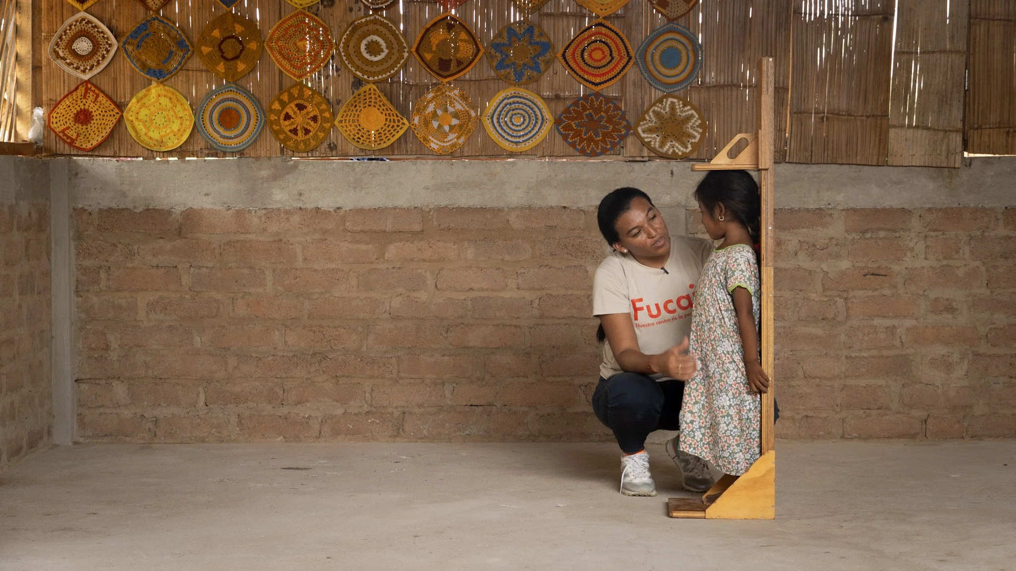 Una mujer y una niña tienen una conversación en un espacio con paredes de ladrillo y un fondo de decoraciones de colores, en un entorno que parece un taller o espacio cultural