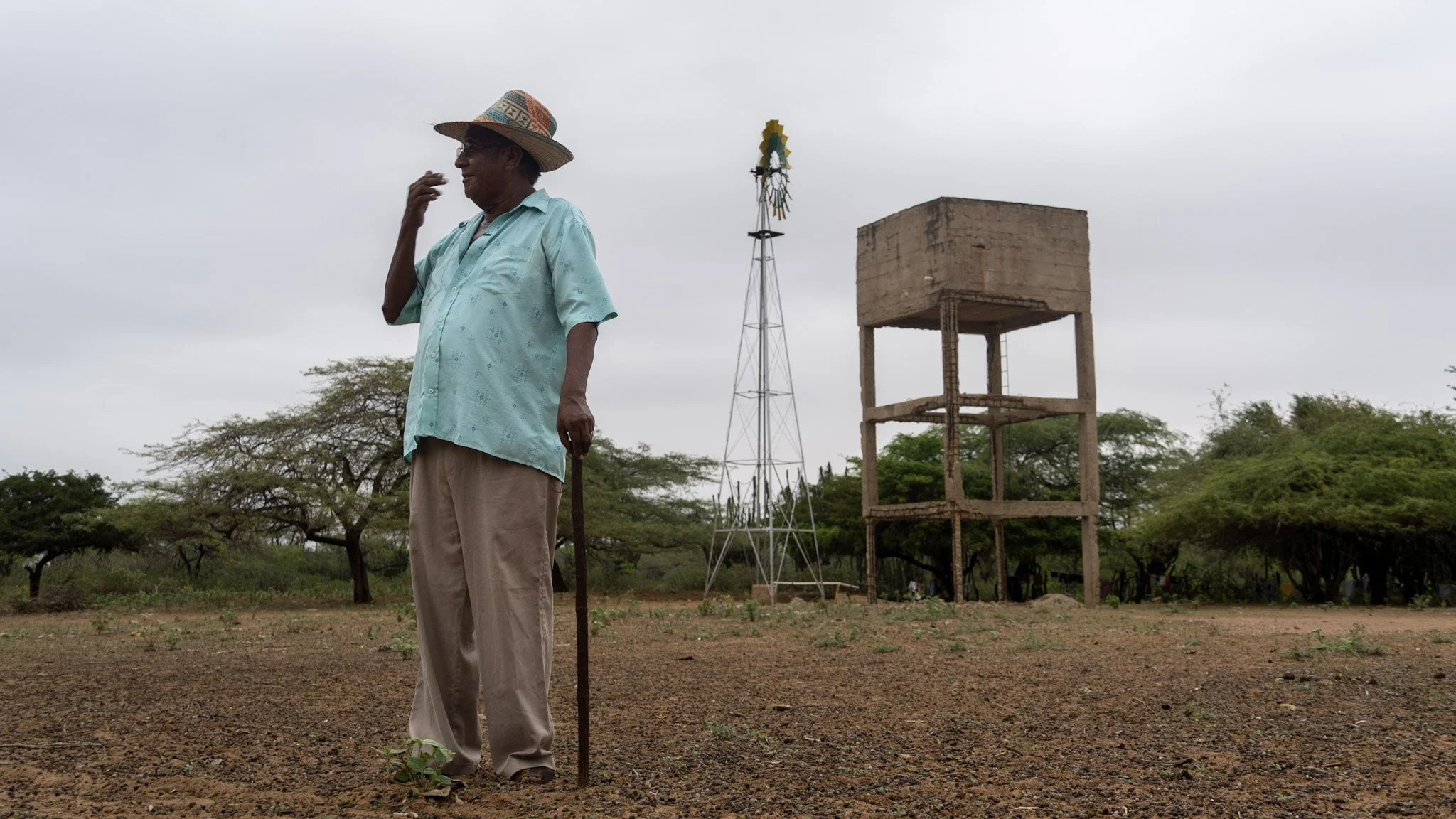 Un hombre mayor con sombrero de paja, camisa azul y pantalones beige, sosteniendo un bastón, en un campo con árboles y estructuras de agua y electricidad, cielo nublado.