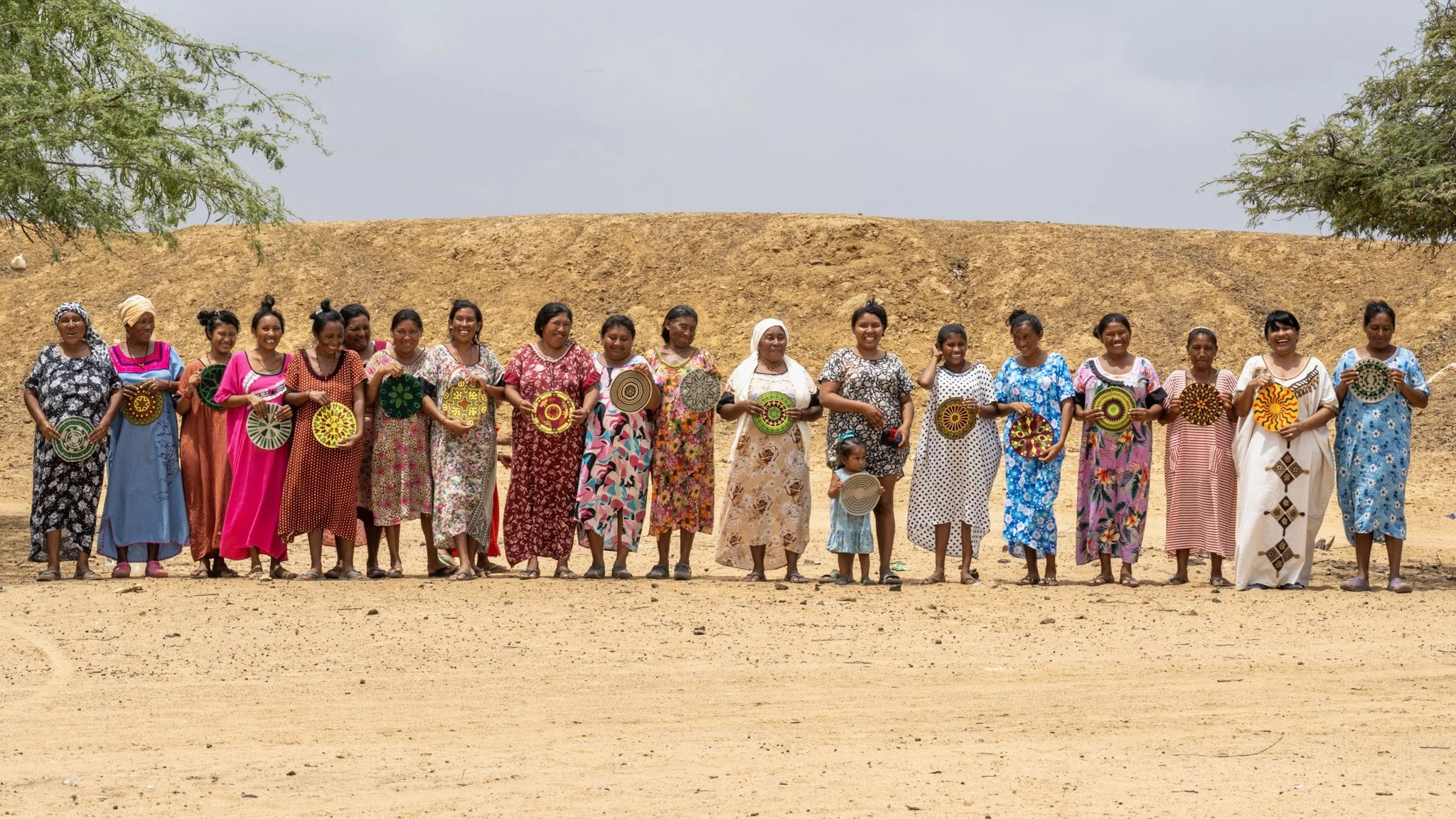 Grupo de mujeres con vestimenta tradicional, sosteniendo artesanías en un campo secó, árboles de fondo y cielo nublado.