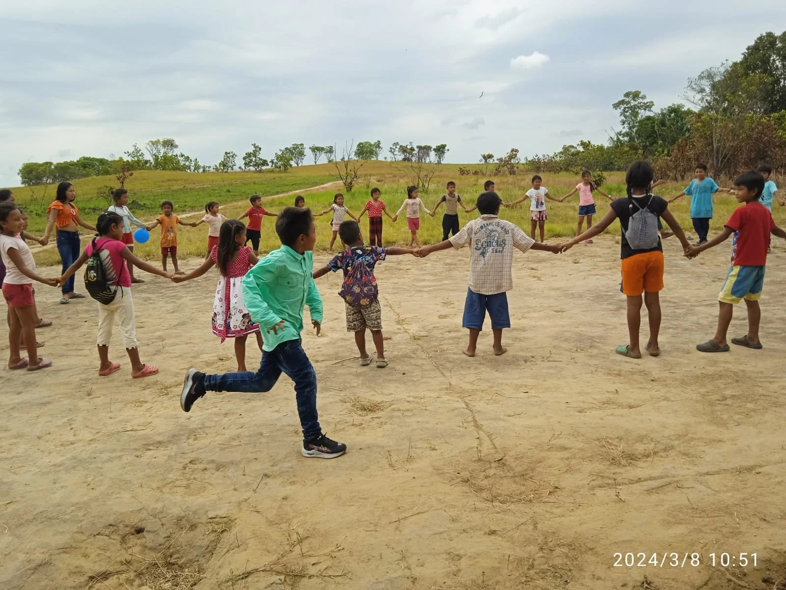 Niños y niñas jugando en círculo al aire libre en un campo, sosteniéndose de las manos y disfrutando del juego, en un día nublado.