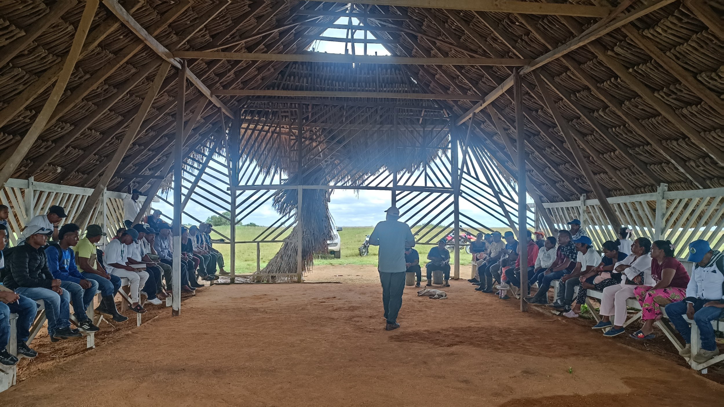 Reunión comunitaria en estructura de madera con techo de palma, personas sentadas en círculo, un hombre hablando en el centro, campo abierto al fondo.