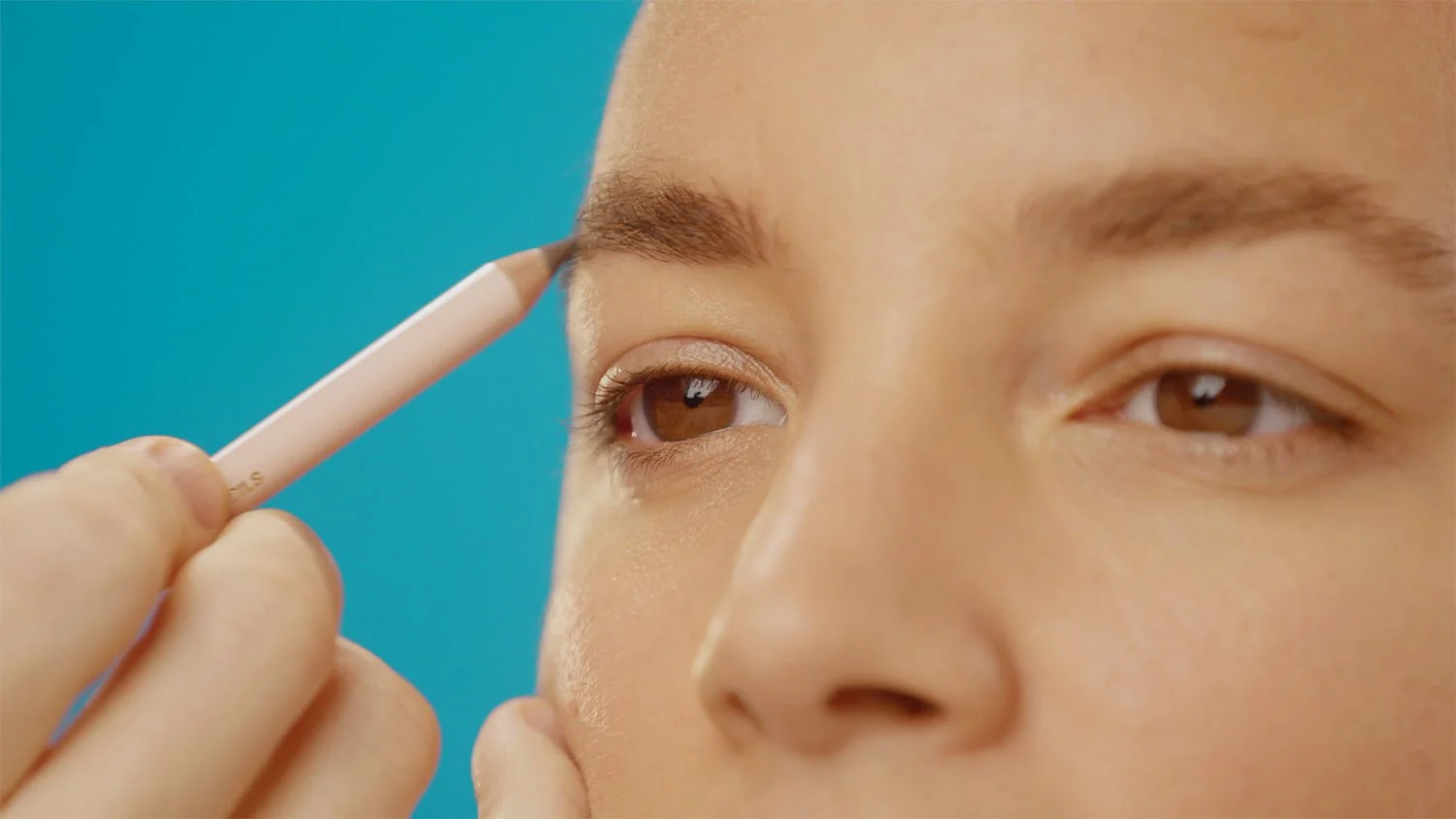 girl getting eyebrows filled in with a pink pencil in front of a blue background