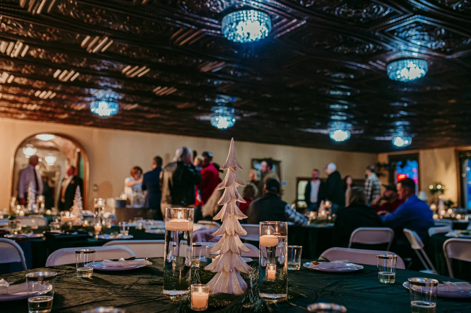 A festive holiday event with tables set for a meal, decorated with small white Christmas trees and candles, and guests mingling and socializing in the background at an indoor venue with decorative ceiling lighting.