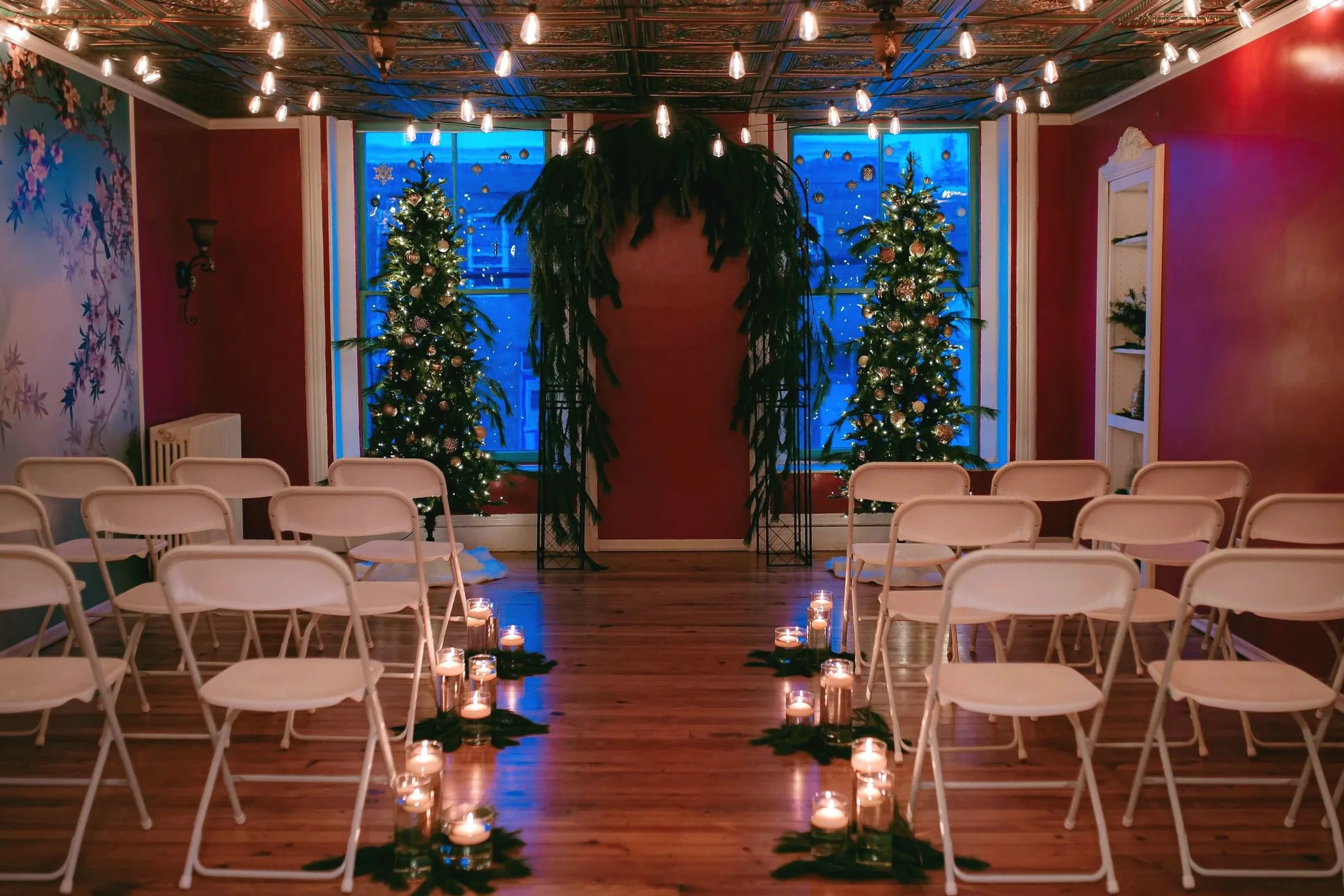 A decorated indoor wedding or event venue with Christmas trees flanking a wooden arch, string lights hanging from the ceiling, and candles lining the aisle. There are white chairs on either side of the aisle and a colorful mural on one wall. Photo Co