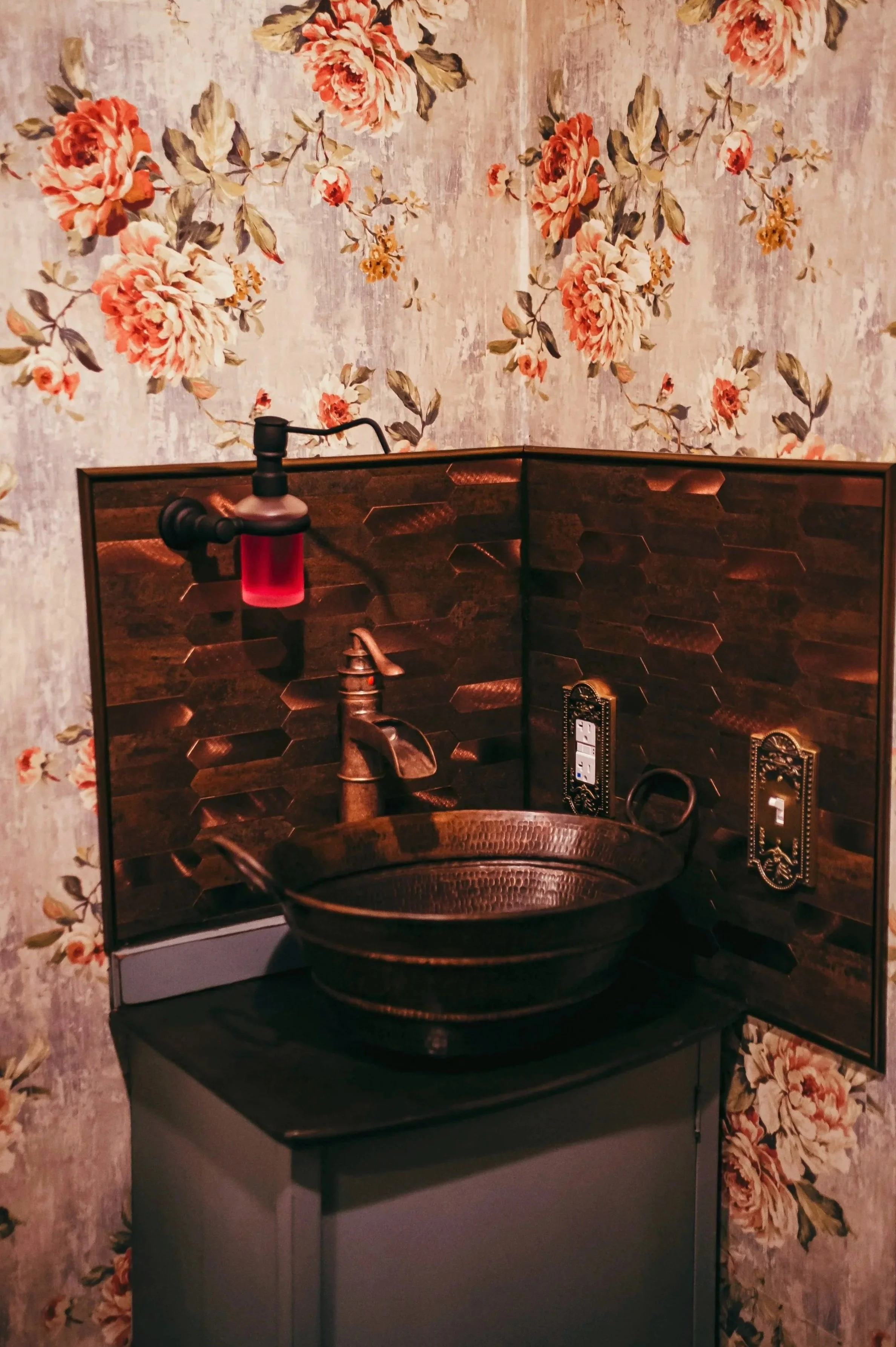 A small bathroom sink with a copper basin, dark wood backsplash, and vintage-style wall fixtures, against floral wallpaper. Photo Courtesy of Shannon Beth Photography.