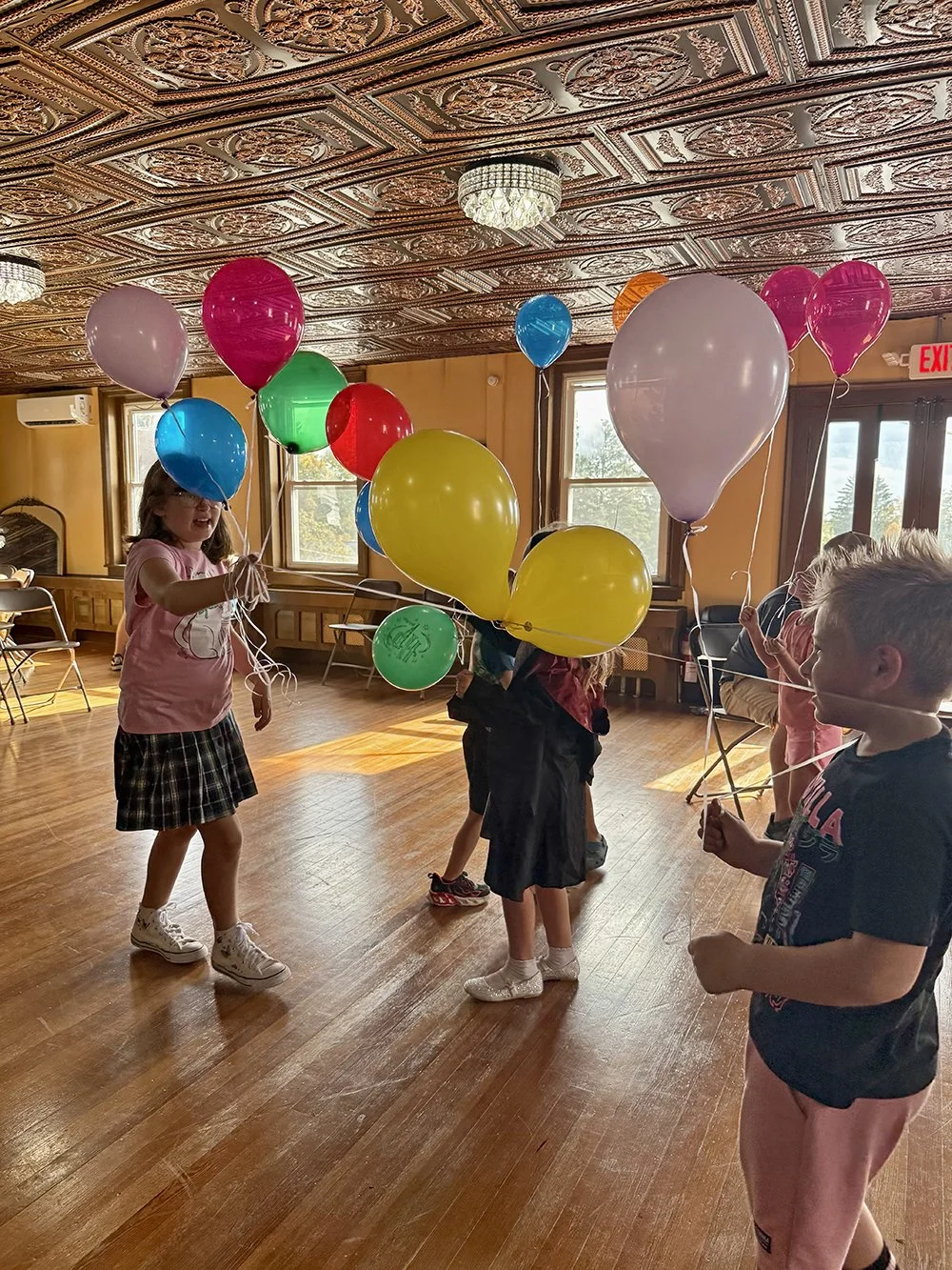 Children holding colorful balloons in a decorated indoor room with wood floors and ornate ceiling panels.