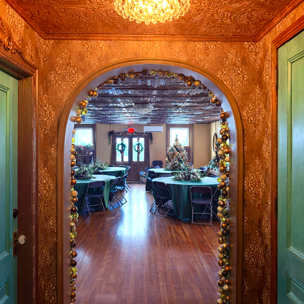 Decorated banquet hall with round tables covered in green tablecloths, floral centerpieces, a decorated Christmas tree, and wreaths on the windows, viewed through a doorway with holiday ornaments.