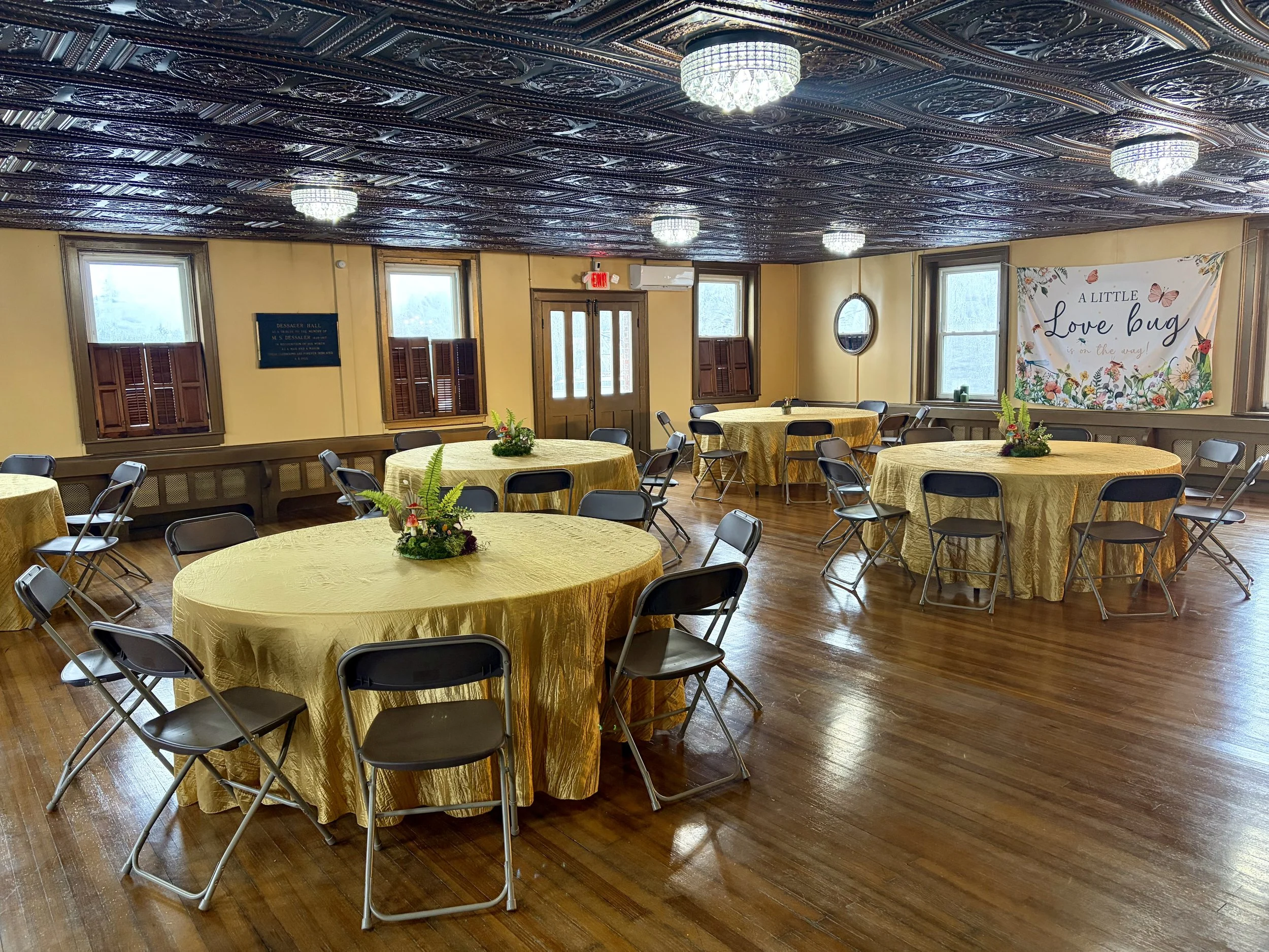 A decorated banquet hall with multiple round tables covered in yellow tablecloths, each with a floral centerpiece, and black folding chairs arranged around them. The hall has wooden floors, windows with wooden shutters, and a decorative ceiling with chandeliers. A banner on the wall reads 'A Little Love Bug'.