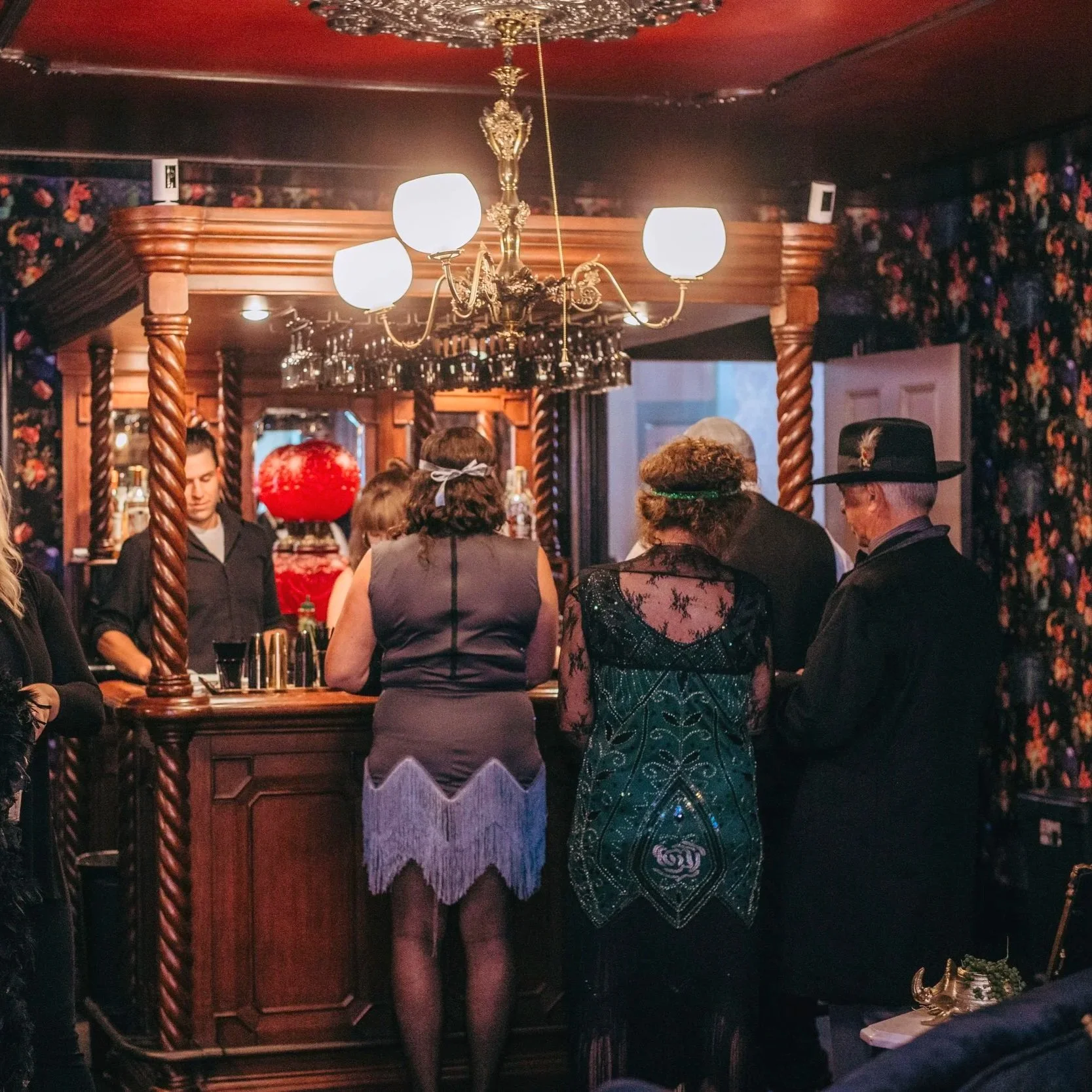 People gathered at a vintage-style bar with wooden details and floral wallpaper, socializing and enjoying drinks under a chandelier with five white globes.