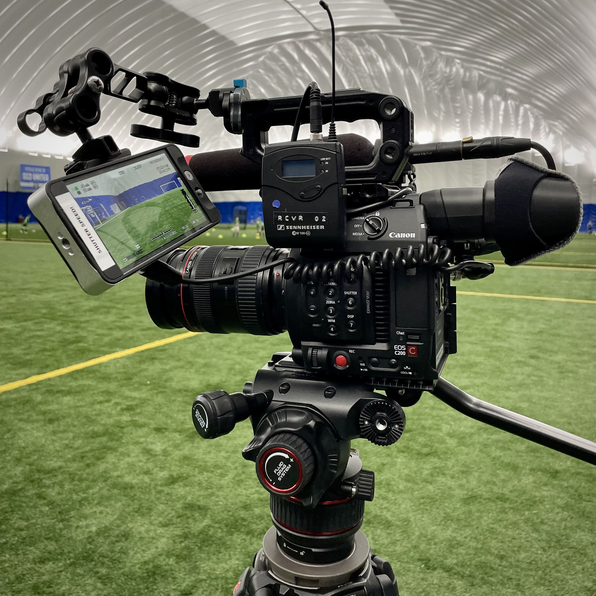 Professional Canon video camera on tripod, featuring Sennheiser microphone receiver, set on indoor sports field with phone mount displaying camera view.