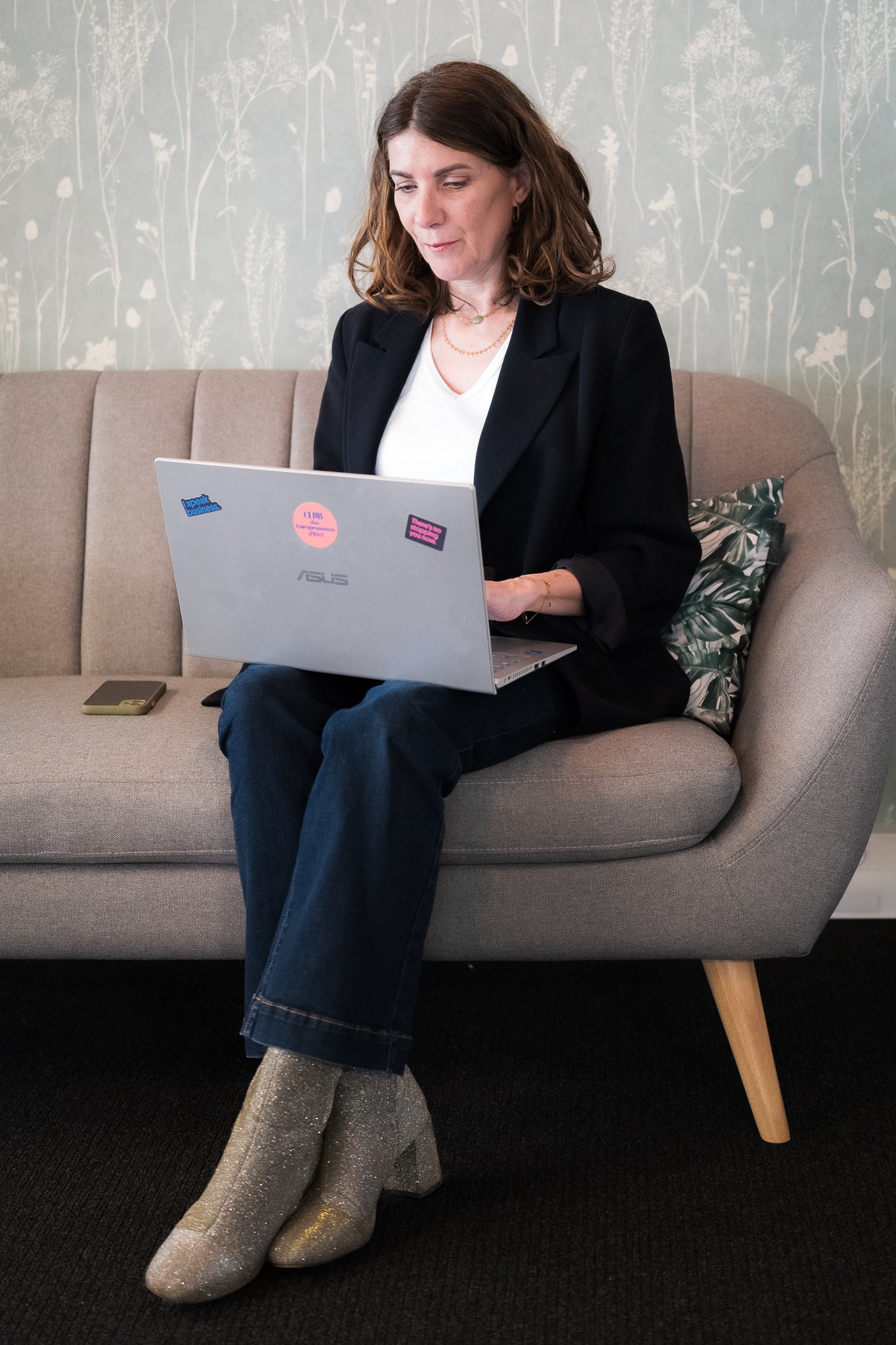 Une femme assise sur un canapé, utilisant un ordinateur portable avec des stickers, portant des vêtements noirs et des chaussettes grises, dans un intérieur décoratif.