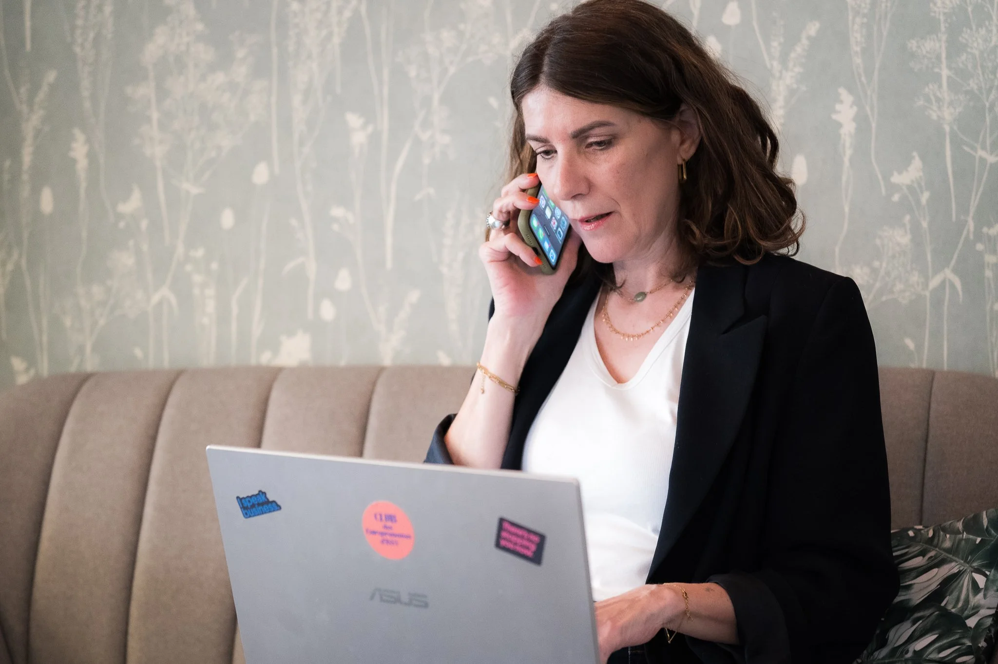 Une femme avec des cheveux bruns, portant une veste noire et un T-shirt blanc, parle au téléphone tout en travaillant sur un ordinateur portable blanc à l'aide d'une souris. Elle est assise sur un canapé beige avec un fond mural décoratif.