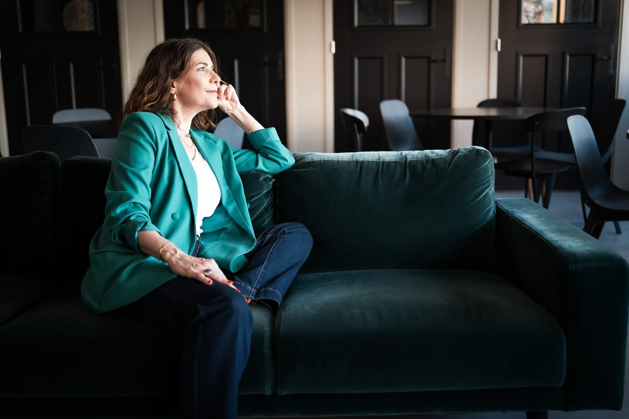 Une femme assise sur un canapé vert, regardant par la fenêtre avec une expression de réflexion. Elle porte une veste verte et des bijoux, dans une pièce avec des murs sombres et des tables en arrière-plan.