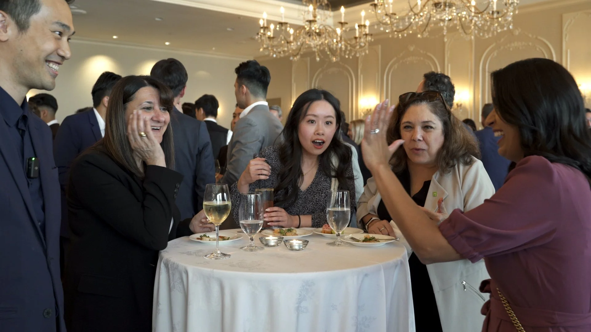 People standing around a table at a social event, laughing and talking, with drinks and appetizers on the table, in a room with chandeliers.