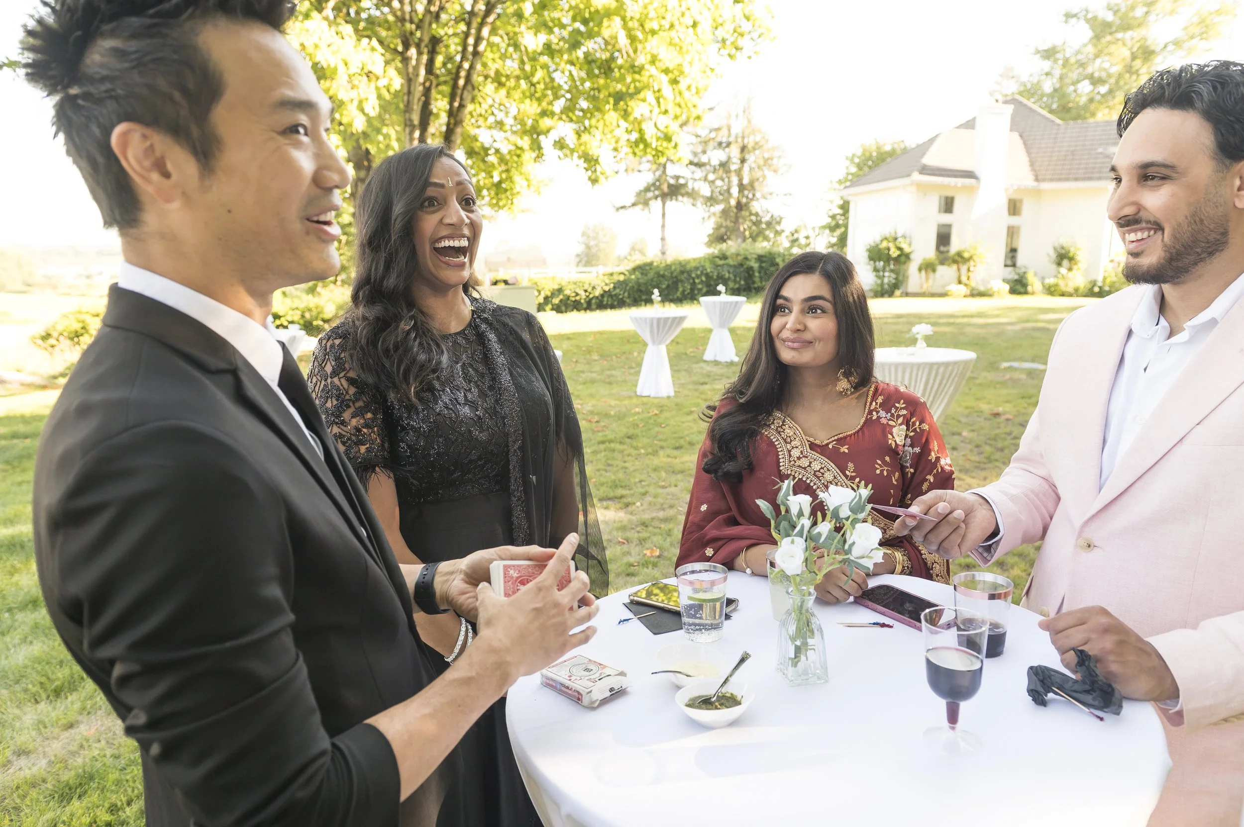 Wedding guests laugh during close-up magic by Kel Ng at Neeraj and Natasha's outdoor Indian wedding cocktail hour in Vancouver