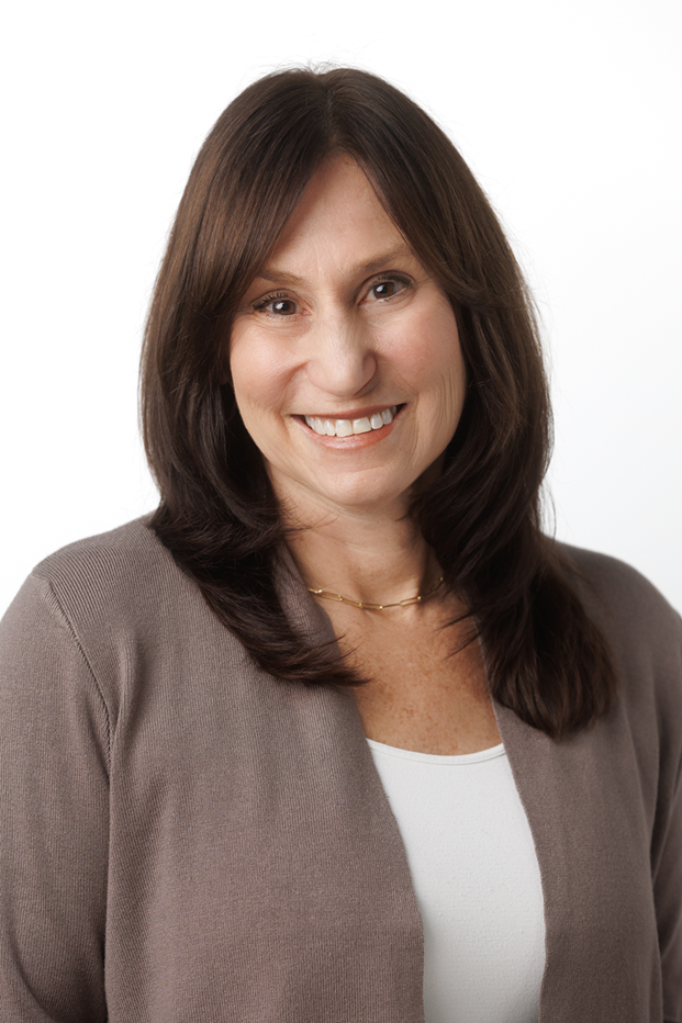 A woman with medium-length brown hair smiling, wearing a brown blazer over a white top, and a delicate gold necklace.