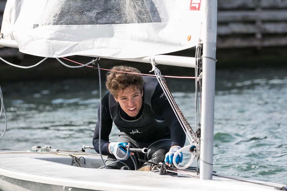 Young male sailor, wearing a gray vest and gloves, intensely handles lines in a small sailboat on the water.