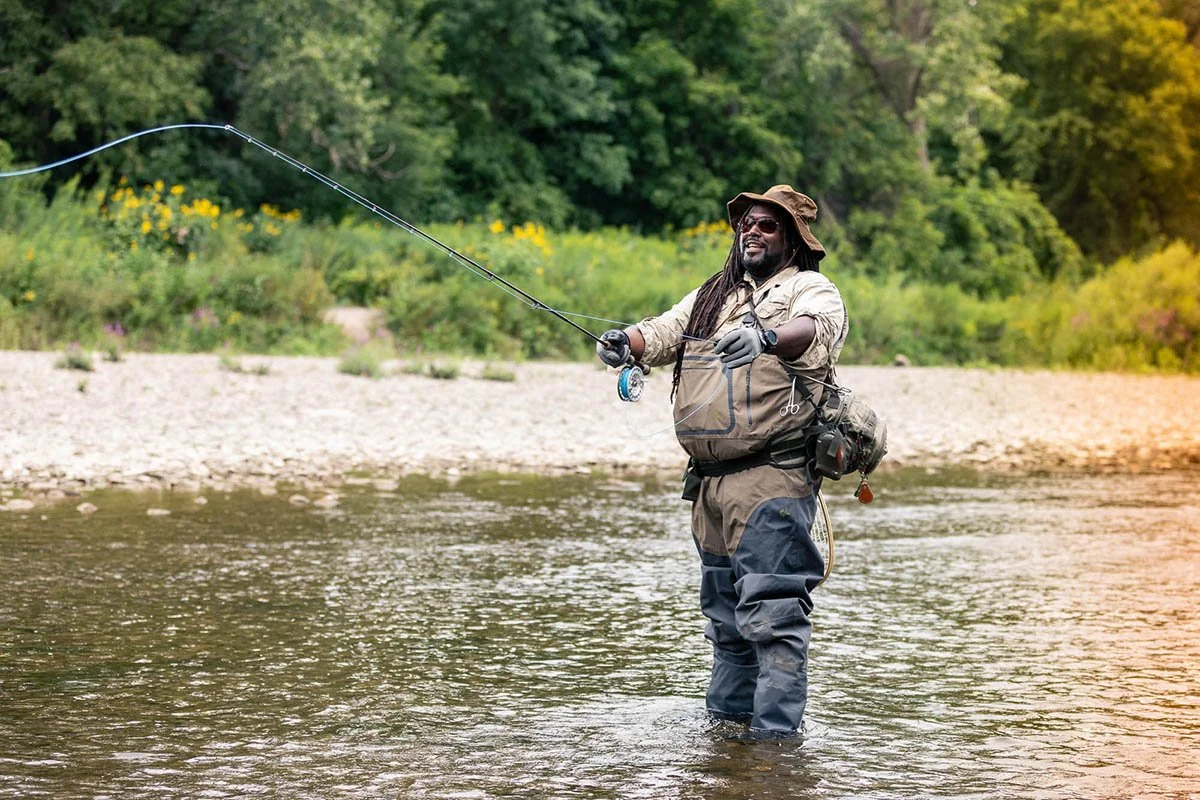 a man wearing gaiters and fishing attire holds fishing rod in a boyd  of water smilig