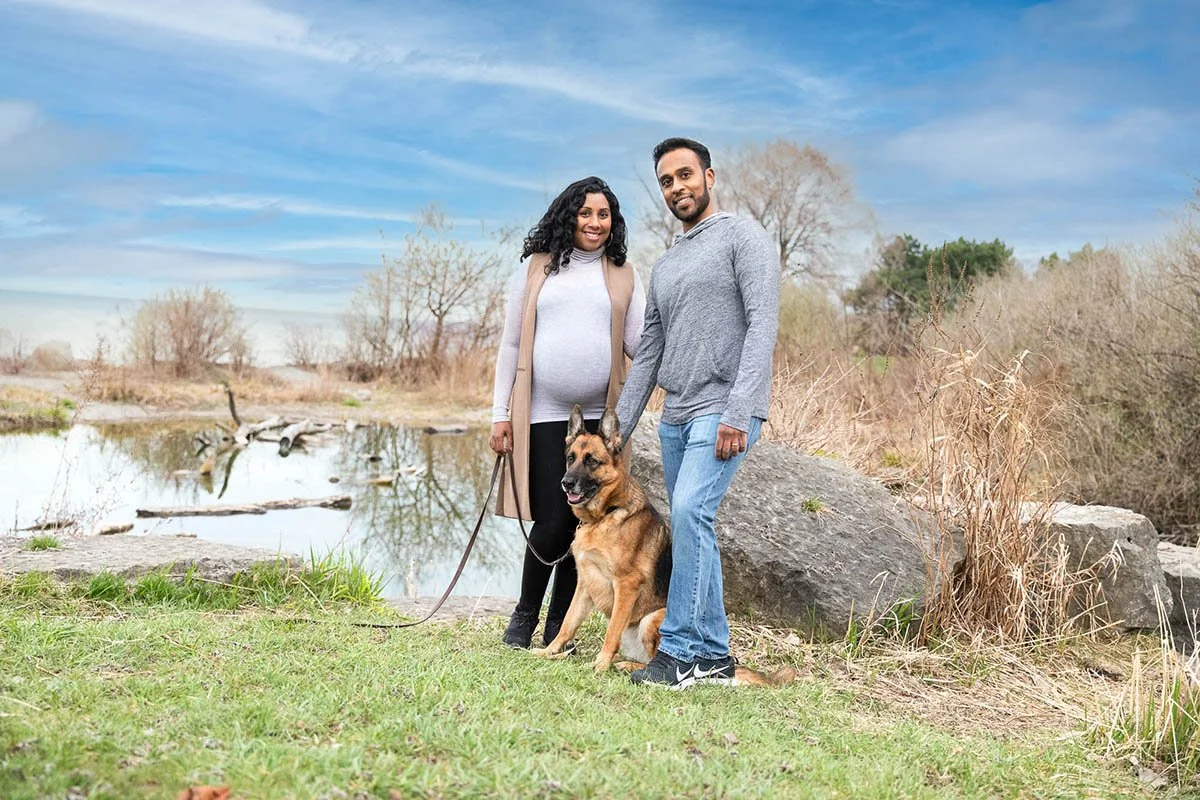 Anne and Jerome pose outdoors with their German Shepherd dog near a pond and a rock.