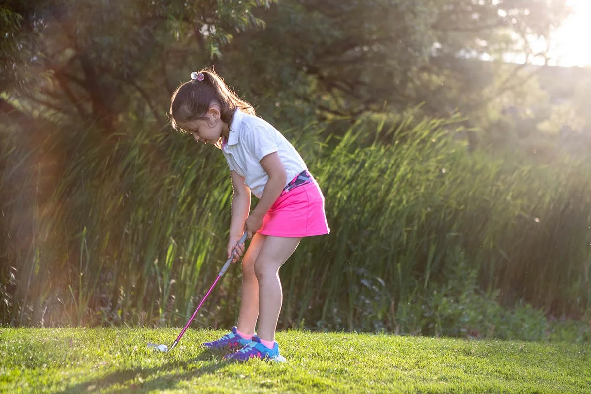 Emma executa uma tacada no campo de golfe com uma expressão concentrada.