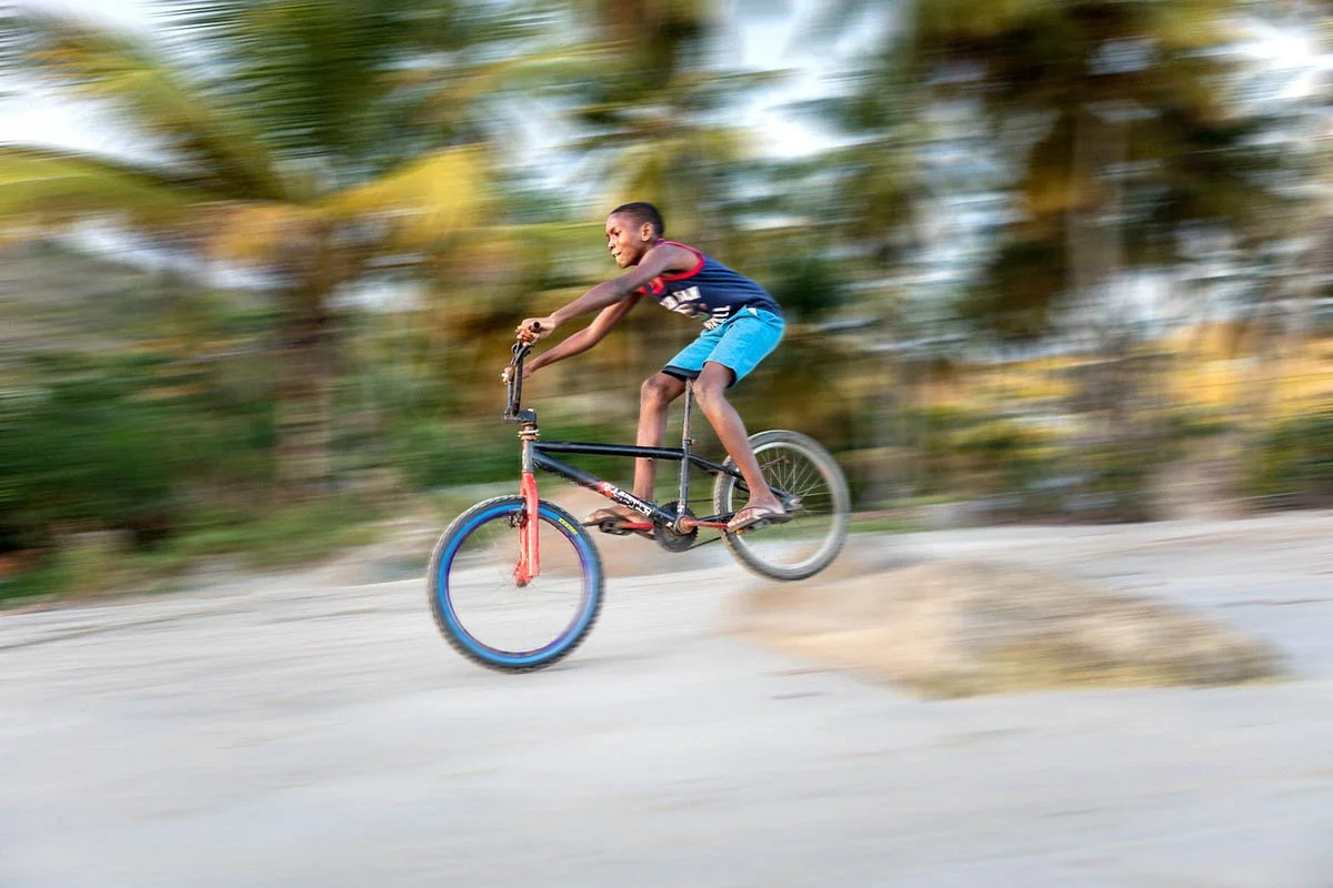 a young saint lucian boy rides his bike on a beach with palm trees blurred in the background