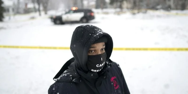 A Black person wearing an I Can't Breath mask with a police car in the background