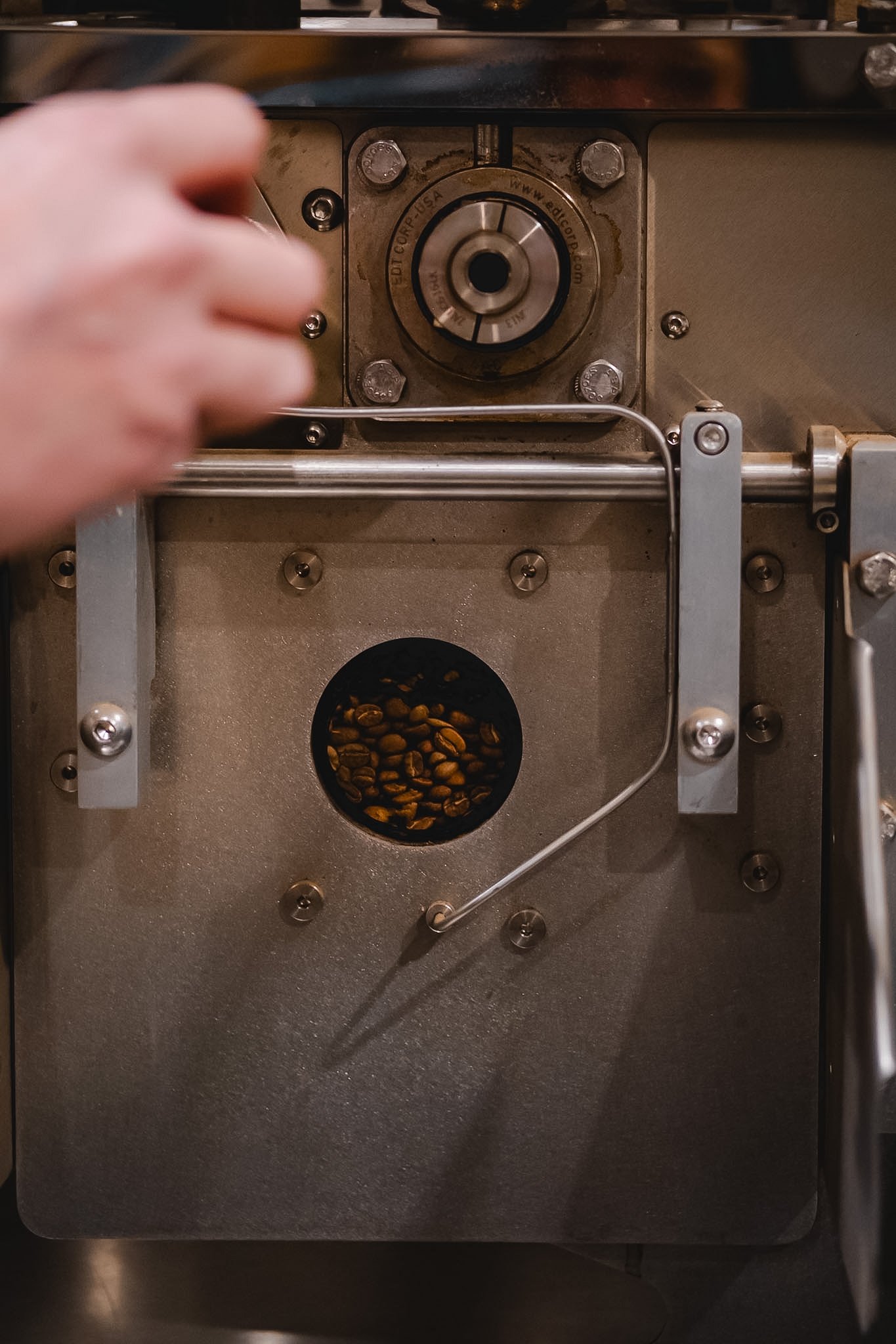 Close up photograph of the front of a loring coffee roaster. Beans are visible in the sightglass. There is a hand holding the coffee trier.