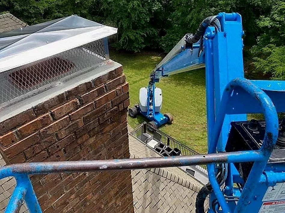 View from a bucket lift showing a blue hydraulic lift extended towards a brick chimney on a rooftop, with a green backyard and trees in the background.