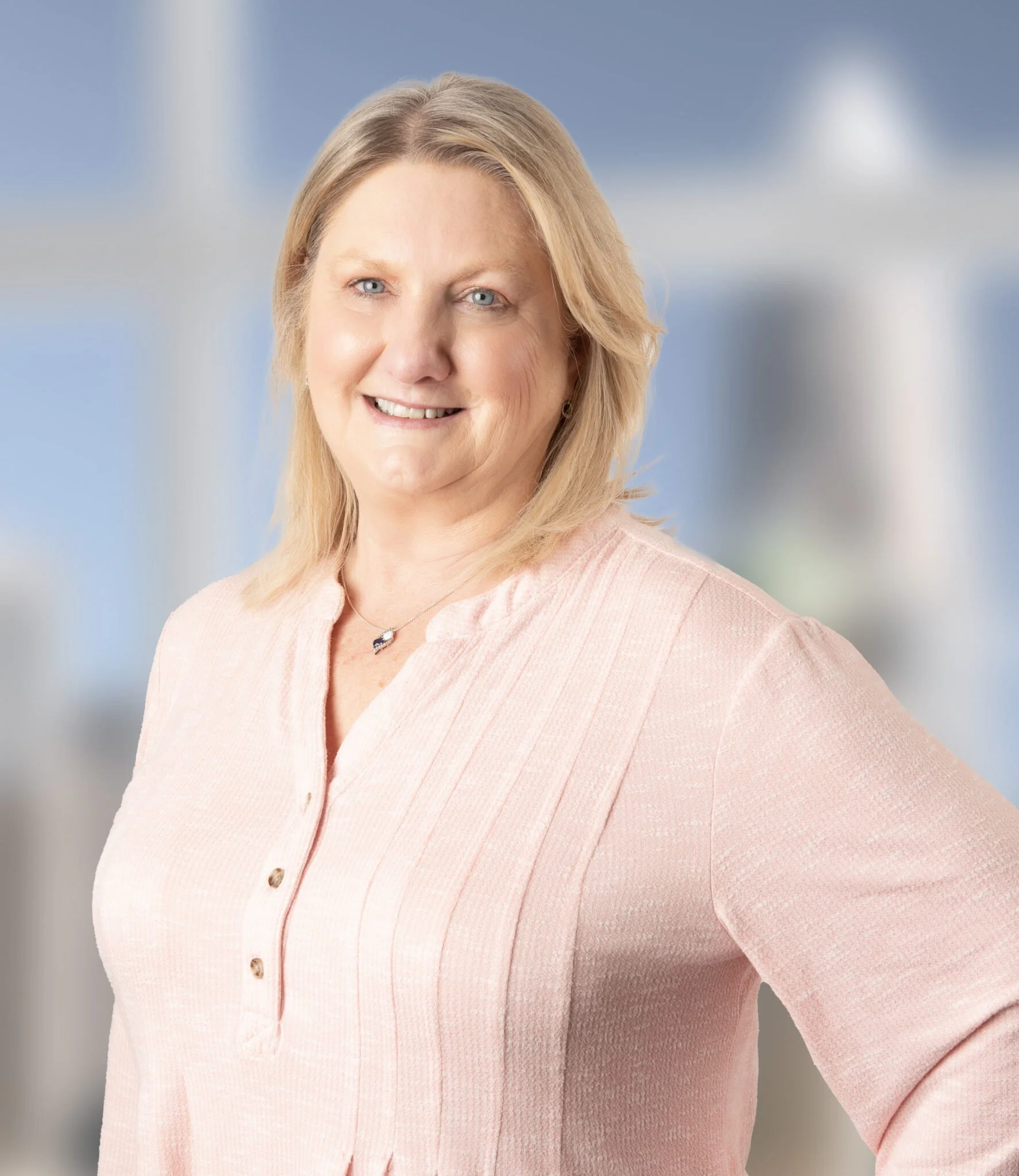 A middle-aged woman with blonde hair, wearing a light pink blouse, smiling and standing with one hand on her hip against a blurred background with a large window.