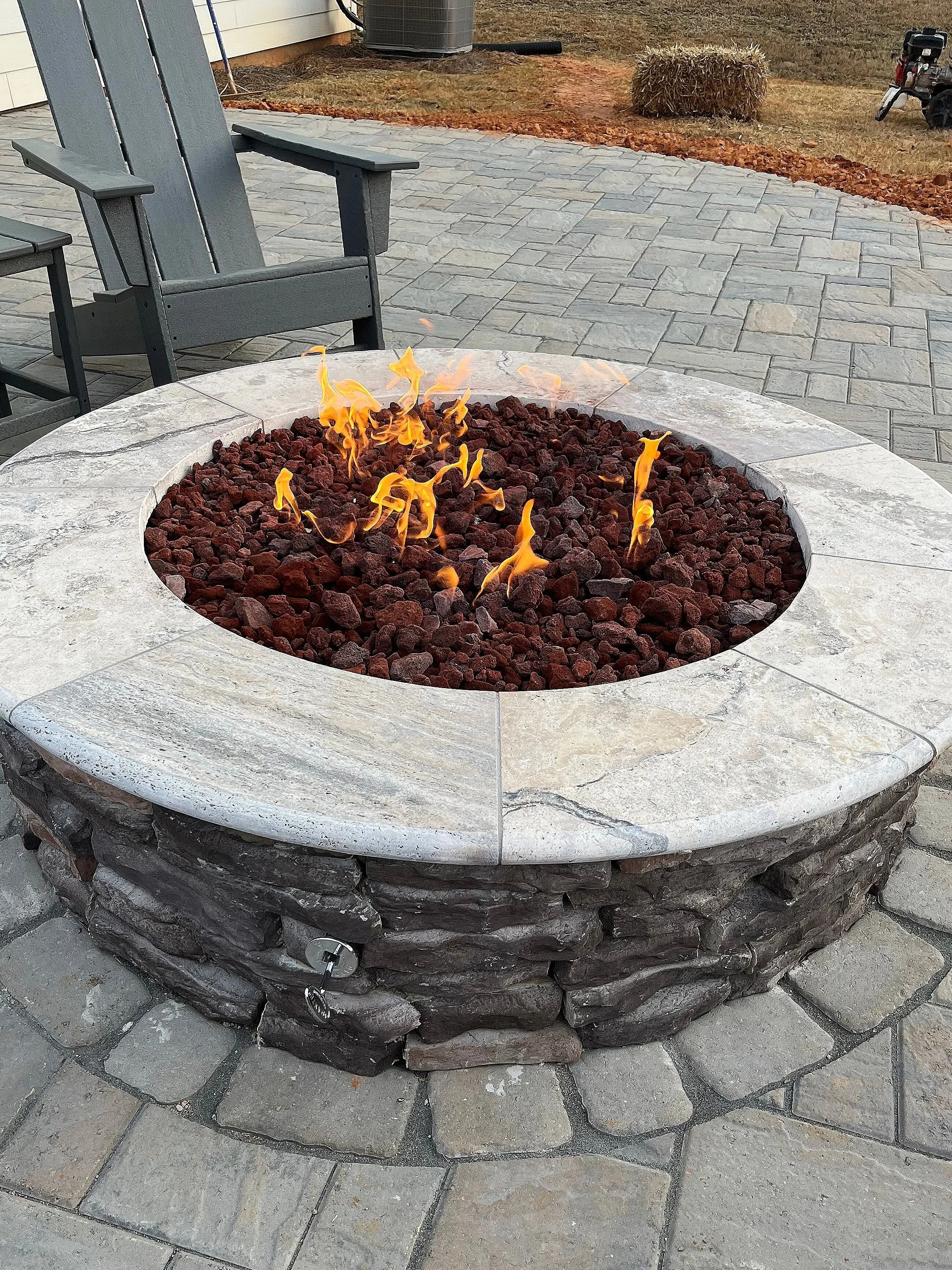 Outdoor fire pit with flames, surrounding stone and brick, on a stone patio with outdoor chairs and a grassy yard.