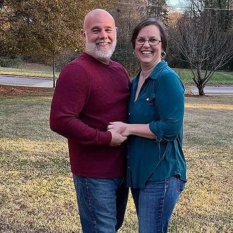 A smiling couple holding hands outdoors in a park with trees and grass.