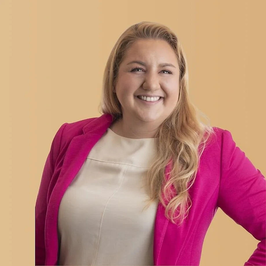 A woman with blonde hair wears a bright pink blazer over a beige top, smiling confidently against a yellow-beige background.