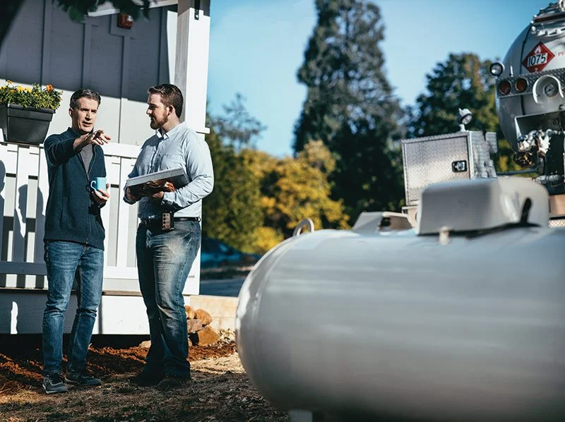 Two men are talking outside a house, one is holding a blue coffee mug and pointing, the other is holding a notebook or clipboard, with a boat and a boat trailer nearby under a blue sky.