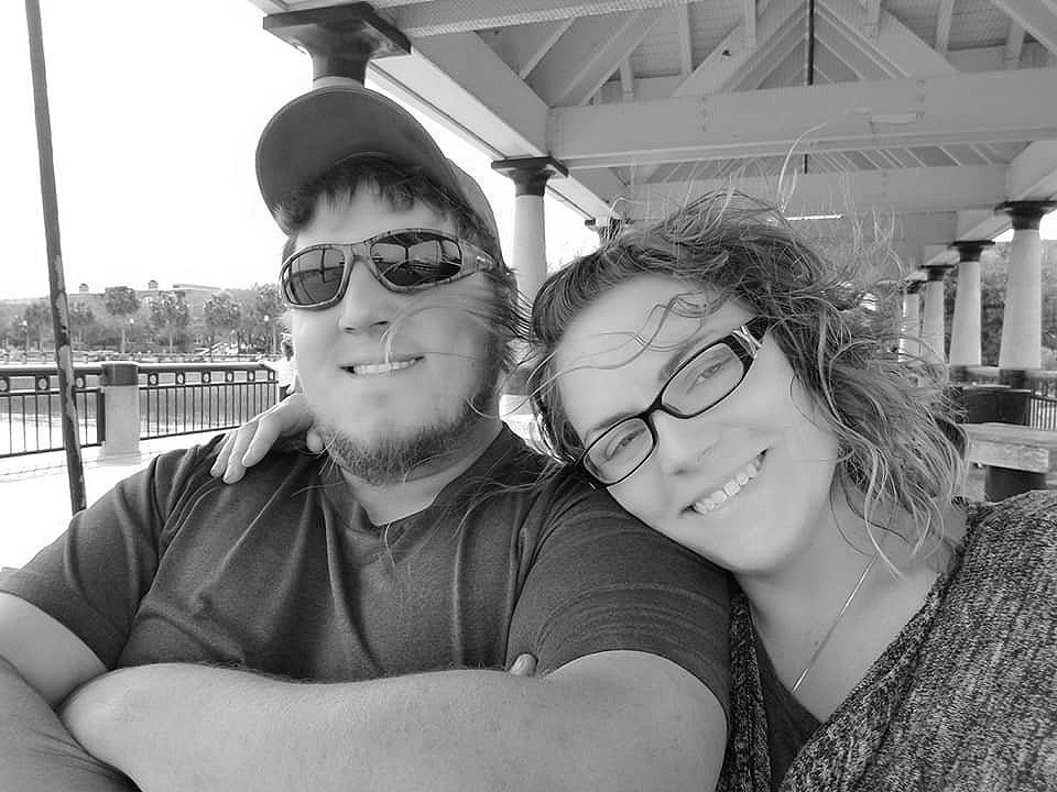 A man and woman are taking a selfie outdoors under a pavilion. The man has a beard, wears sunglasses and a cap, and is smiling. The woman has curly hair, wears glasses, and is smiling with her eyes closed.