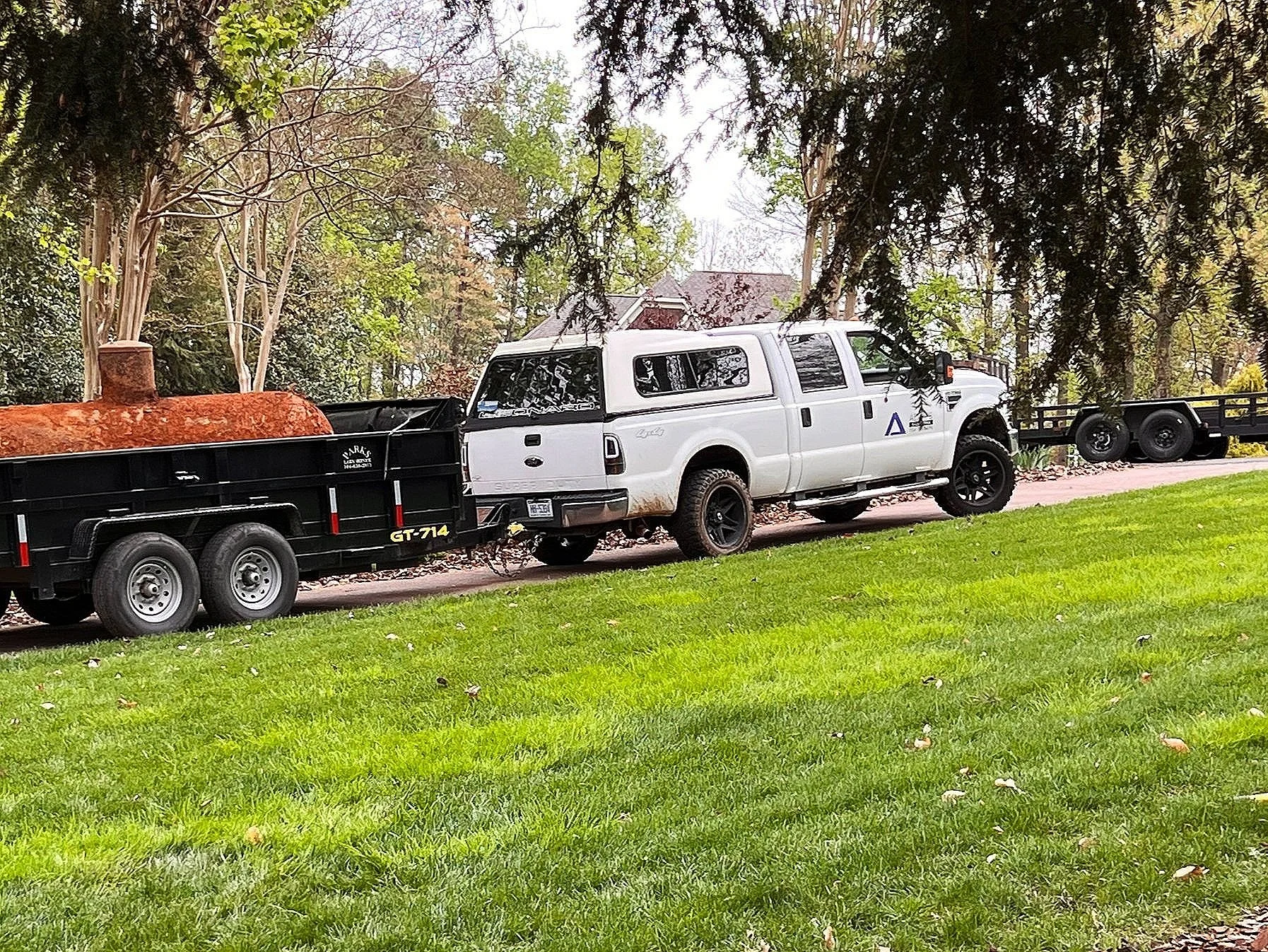 White pickup truck with a camper shell pulling a black trailer and a second black trailer behind it, parked on grass and driveway under trees.
