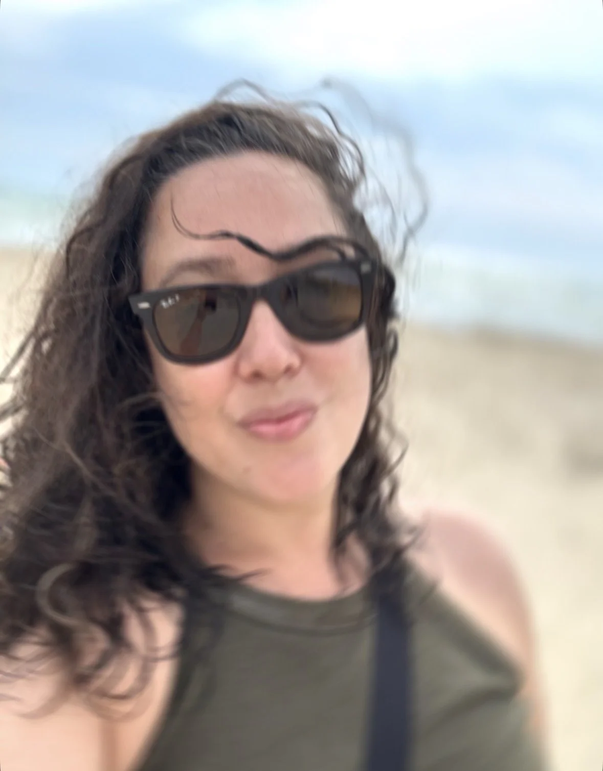 A woman with curly hair wearing sunglasses and a sleeveless top, smiling at the beach.