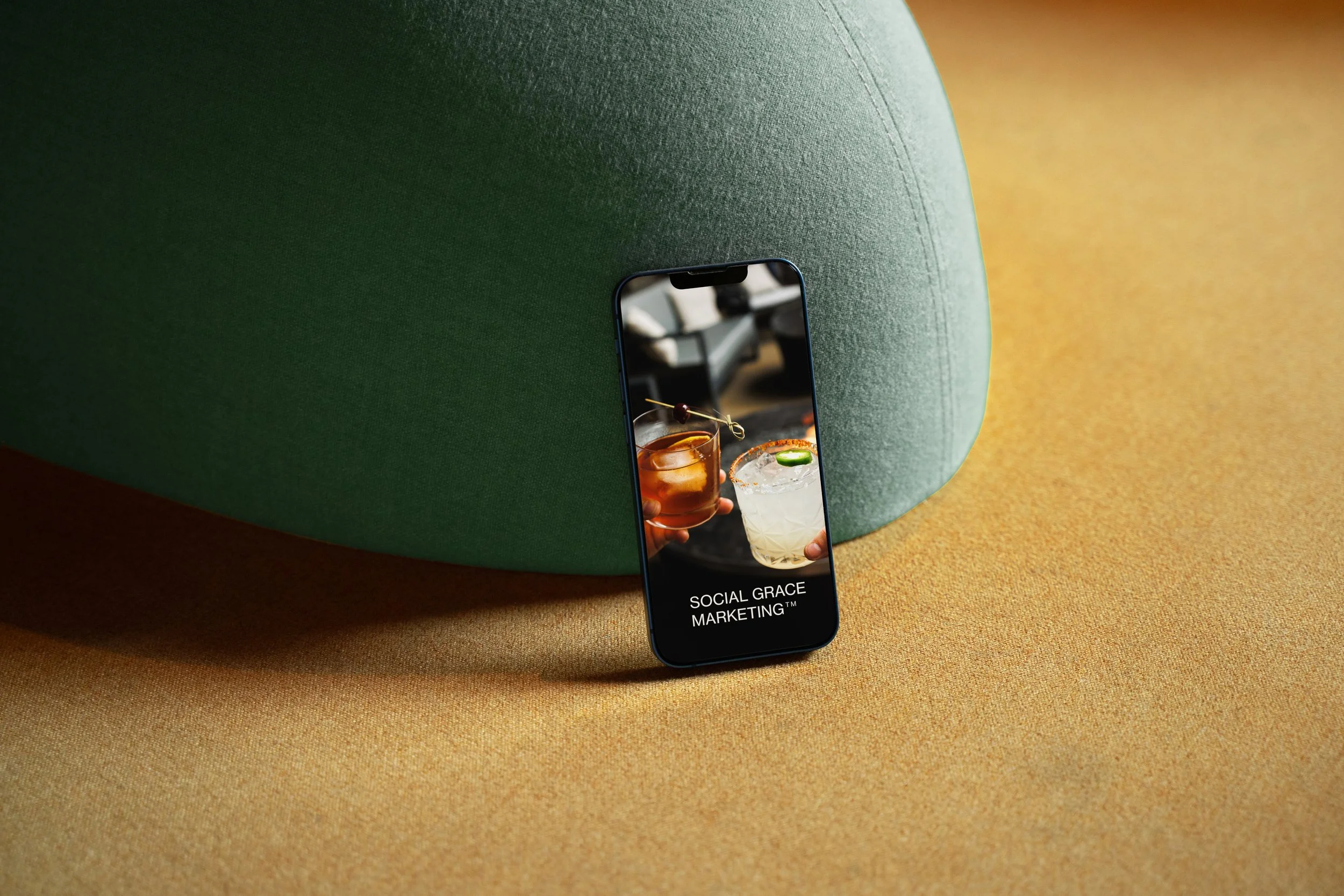 Smartphone leaning against a large green fabric-covered chair with a beige surface. The phone screen displays two cocktails being clinked together, with the text "Social Grace Marketing™" at the bottom.