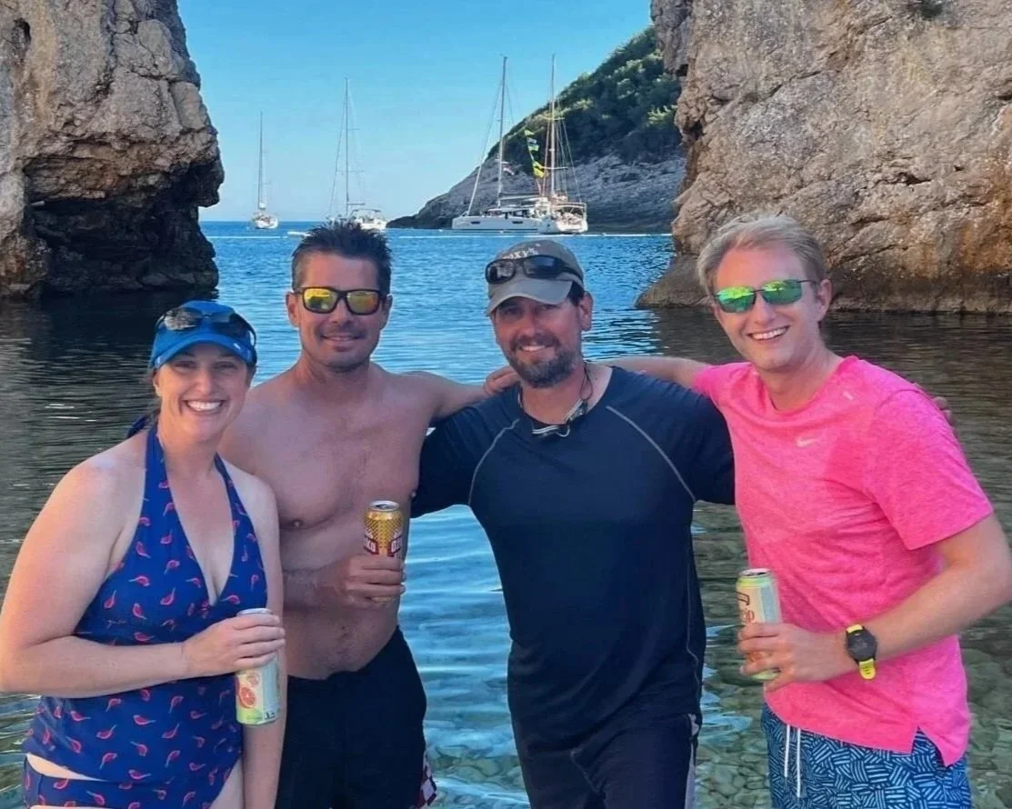 Four people standing in the water by a rocky coastline, smiling and holding drinks, with sailboats and a small island in the background on a sunny day.