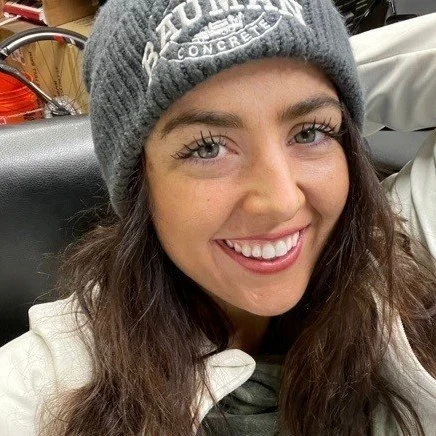 A woman with long, dark hair wearing a gray knit beanie and a white jacket, smiling at the camera.