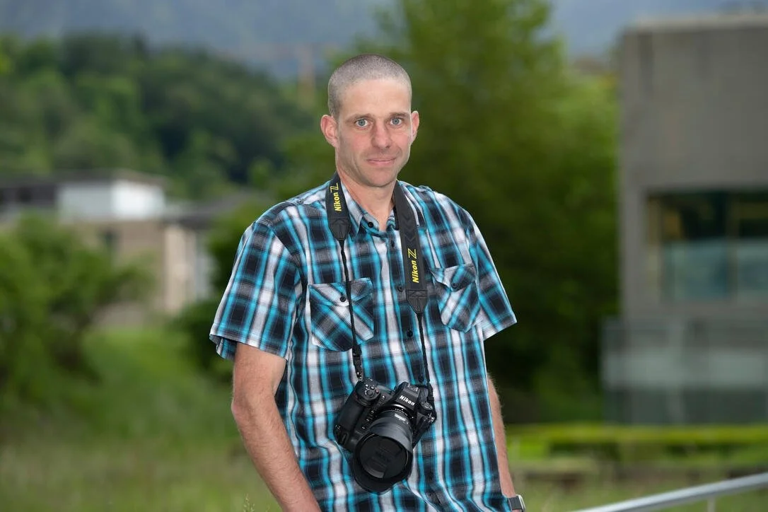 Mann mit kariertem Hemd und Kamera um den Hals, im Freien mit unscharfem Hintergrund.