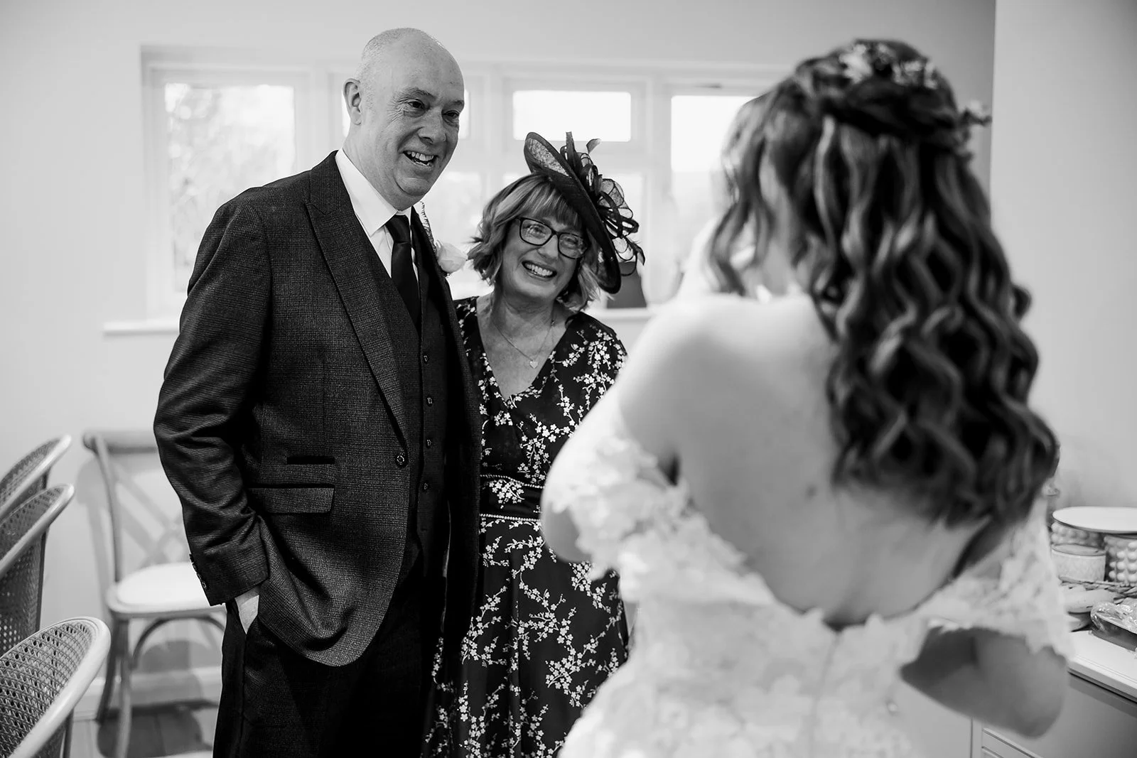 A black and white photo of a wedding moment, with a bride facing her parents, a man and a woman, who are smiling at her. The man is wearing a suit, and the woman is wearing a floral dress and a large hat. The bride has long, curly hair and is in a la