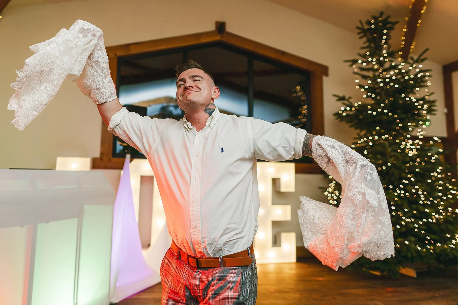 A man in a white shirt and patterned pants with a belt, wearing removable wedding dress sleeves, dancing in front of a decorated Christmas tree and illuminated love letters at a Christmas wedding
