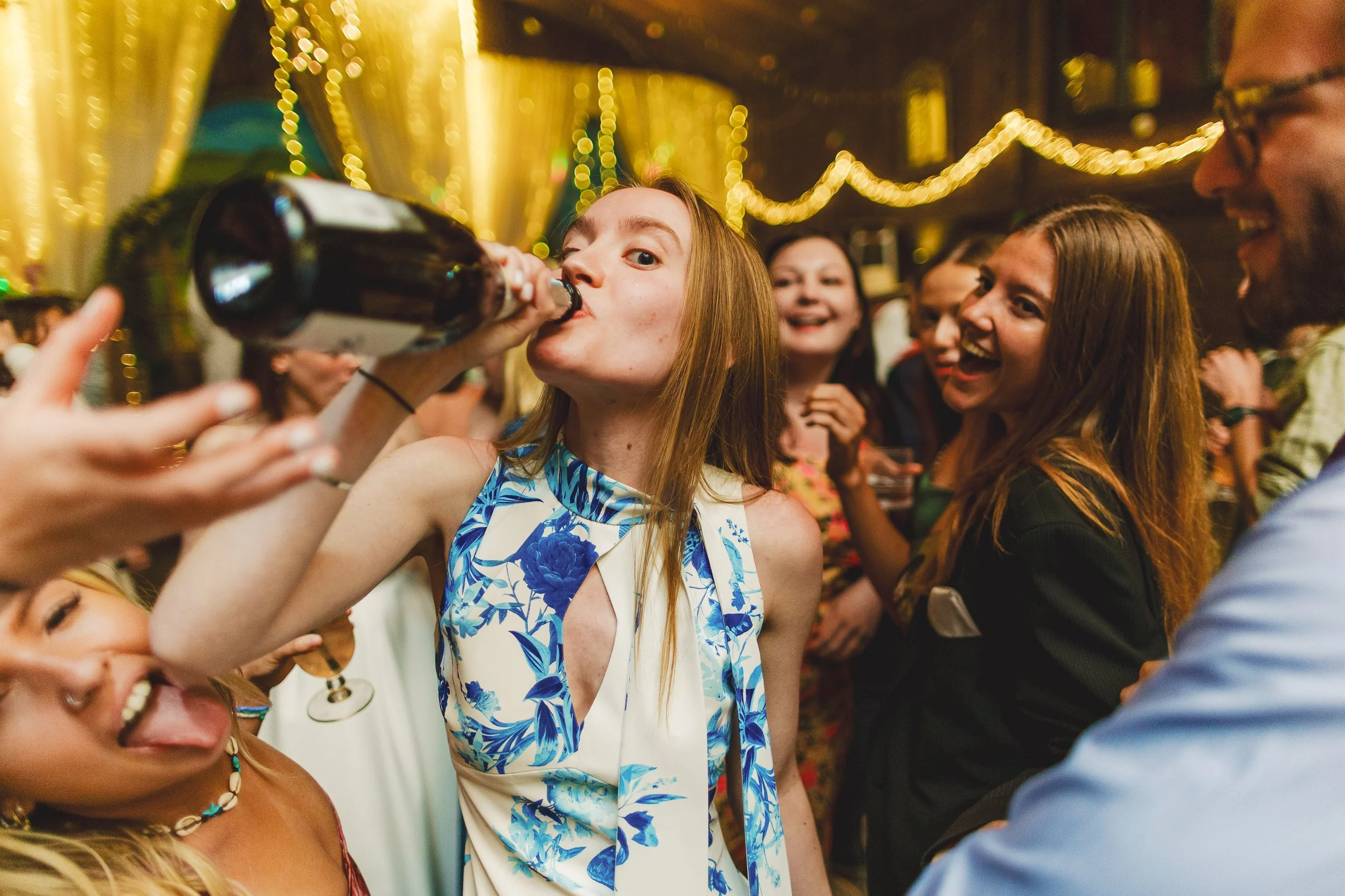 Group of people celebrating at a barn wedding with warm lighting and string lights, with one woman drinking from a bottle while others laugh and enjoy.