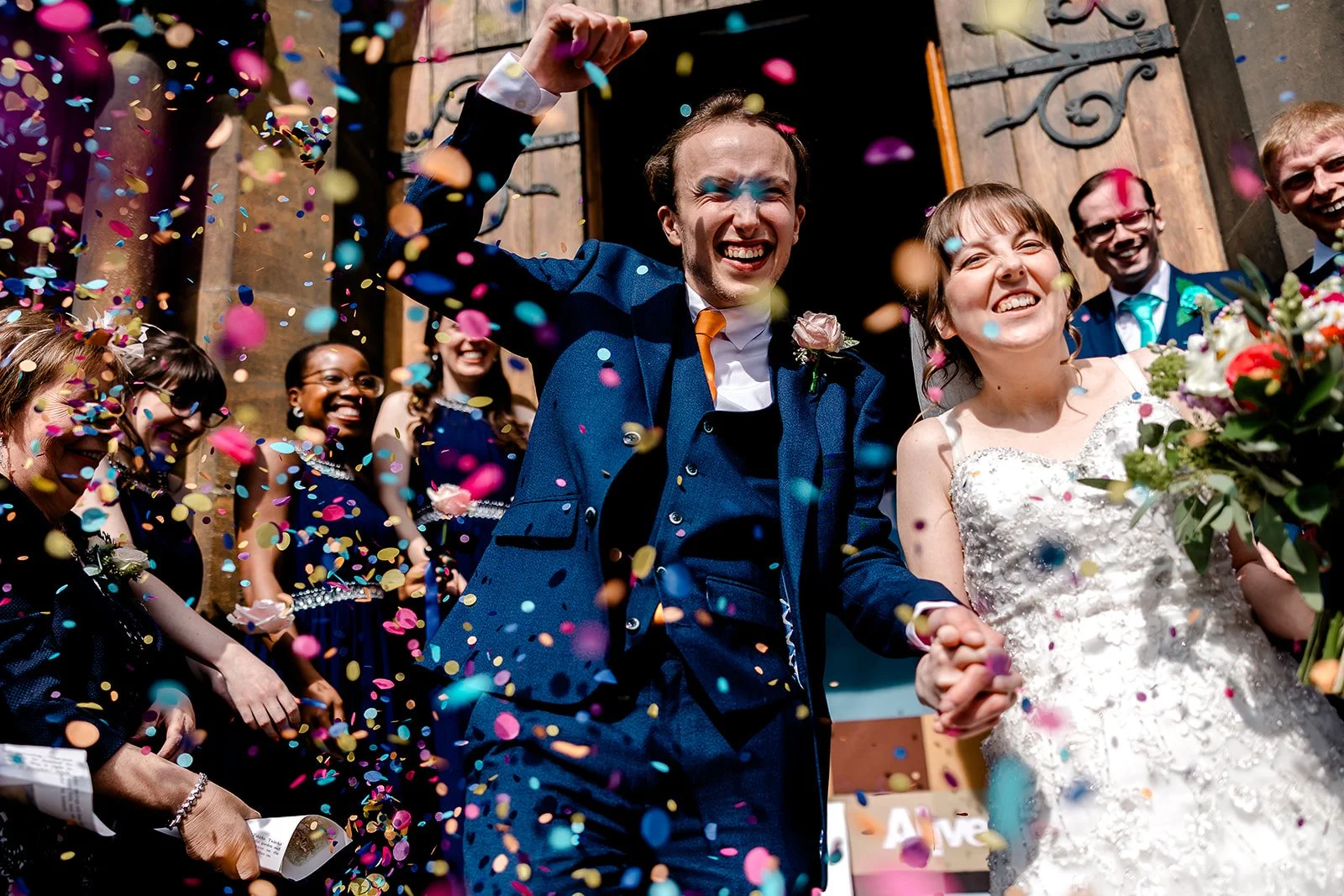 A couple dressed in wedding attire holding hands and smiling amidst colorful confetti raining down during their wedding celebration outside a church entrance.