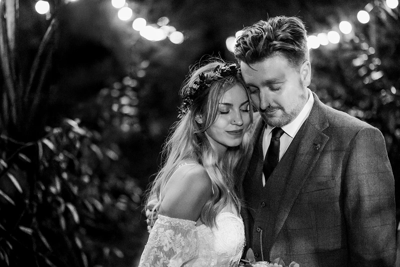 A black-and-white photo of a bride and groom standing close together with eyes closed, sharing an intimate moment outdoors at night, with blurred lights in the background.