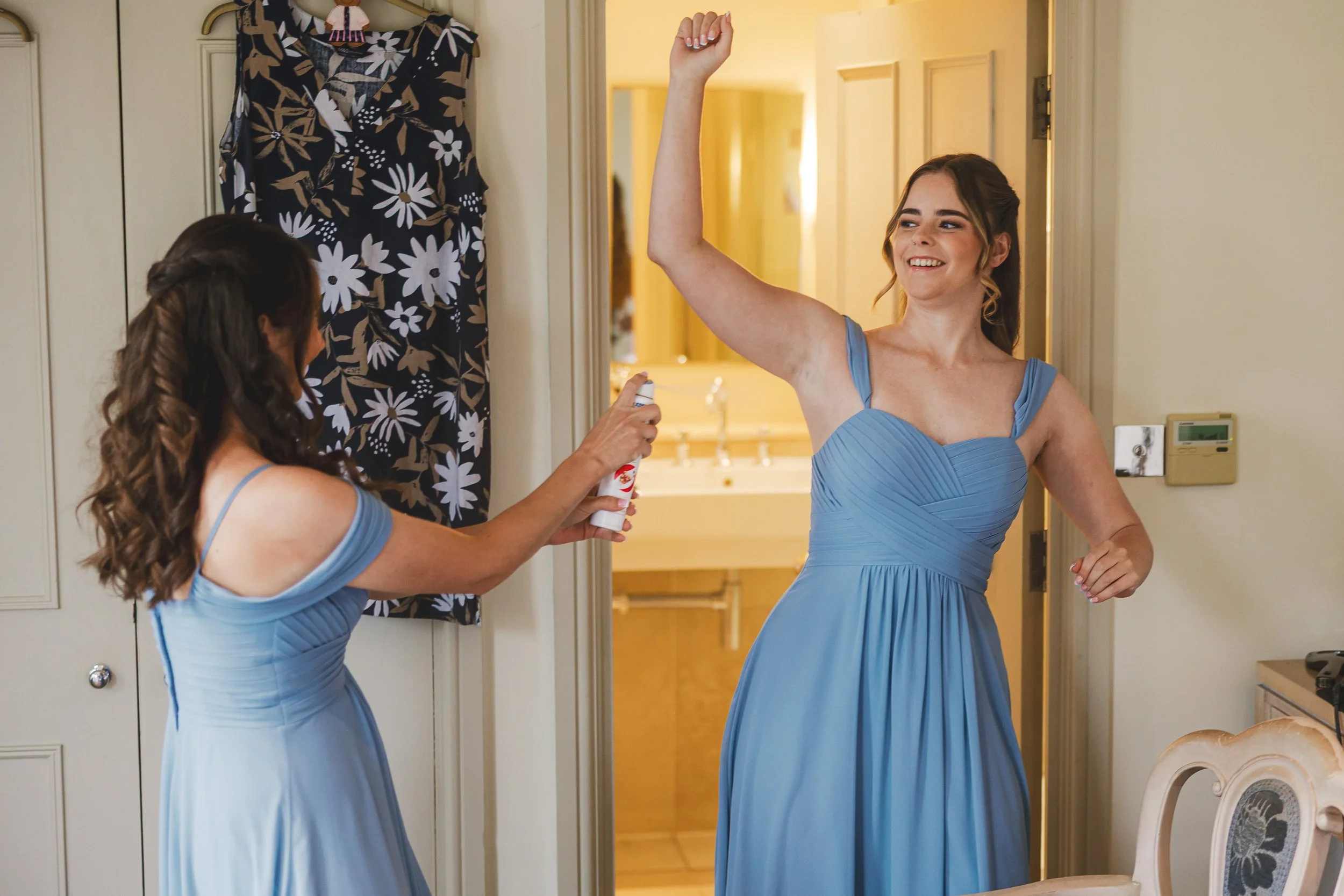 A bridesmaid in a blue dress is smiling and raising her arm as another bridesmaid in a matching blue dress sprays her with a can of deodorant.