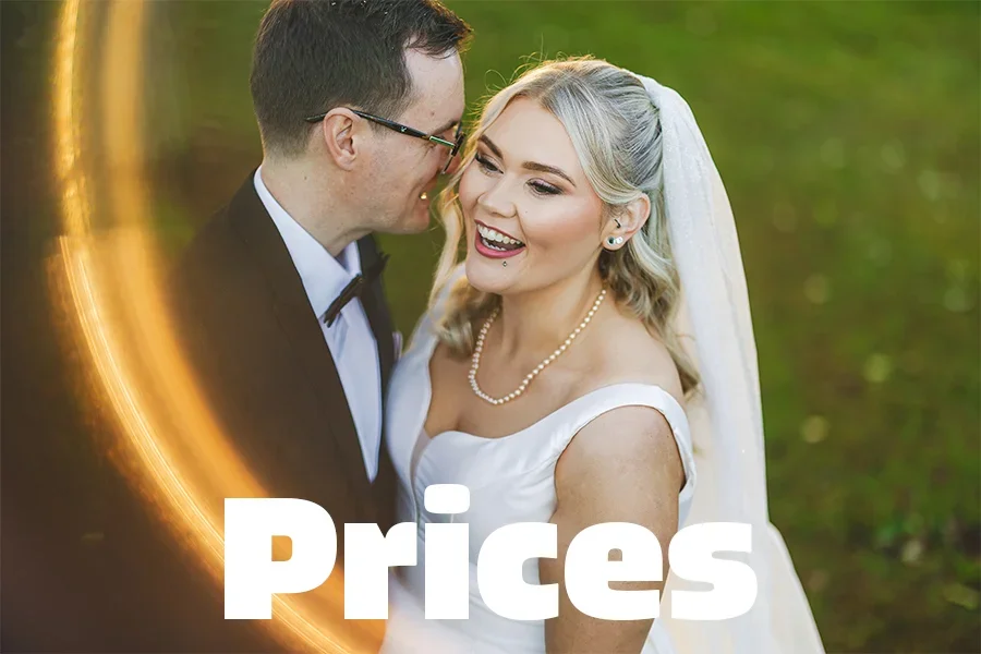 Wedding photo of a smiling bride with blonde hair in a white dress and pearl necklace, and a groom in glasses and a tuxedo, outdoors with green background.