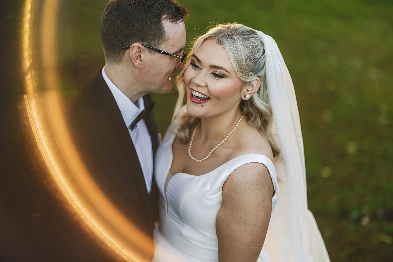 A newlywed couple with the groom leaning in to the bride, smiling outdoors with a blurred green background and ring of fire.