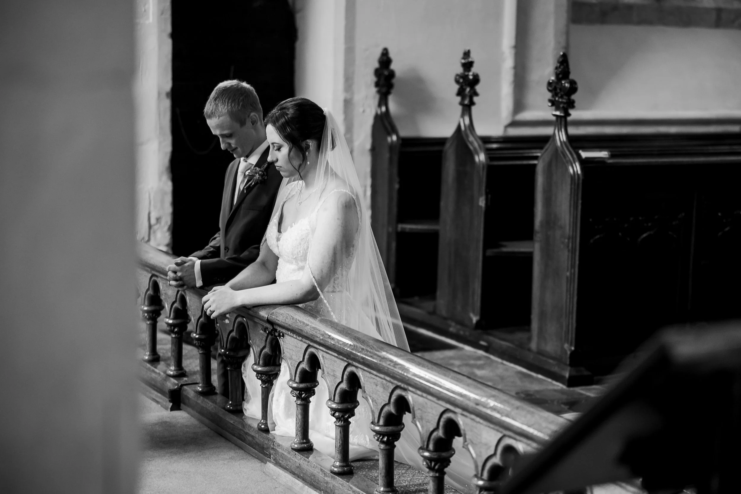 A black-and-white photograph of a bride and groom kneeling at the altar during a wedding ceremony in a church, with their heads bowed and hands clasped.